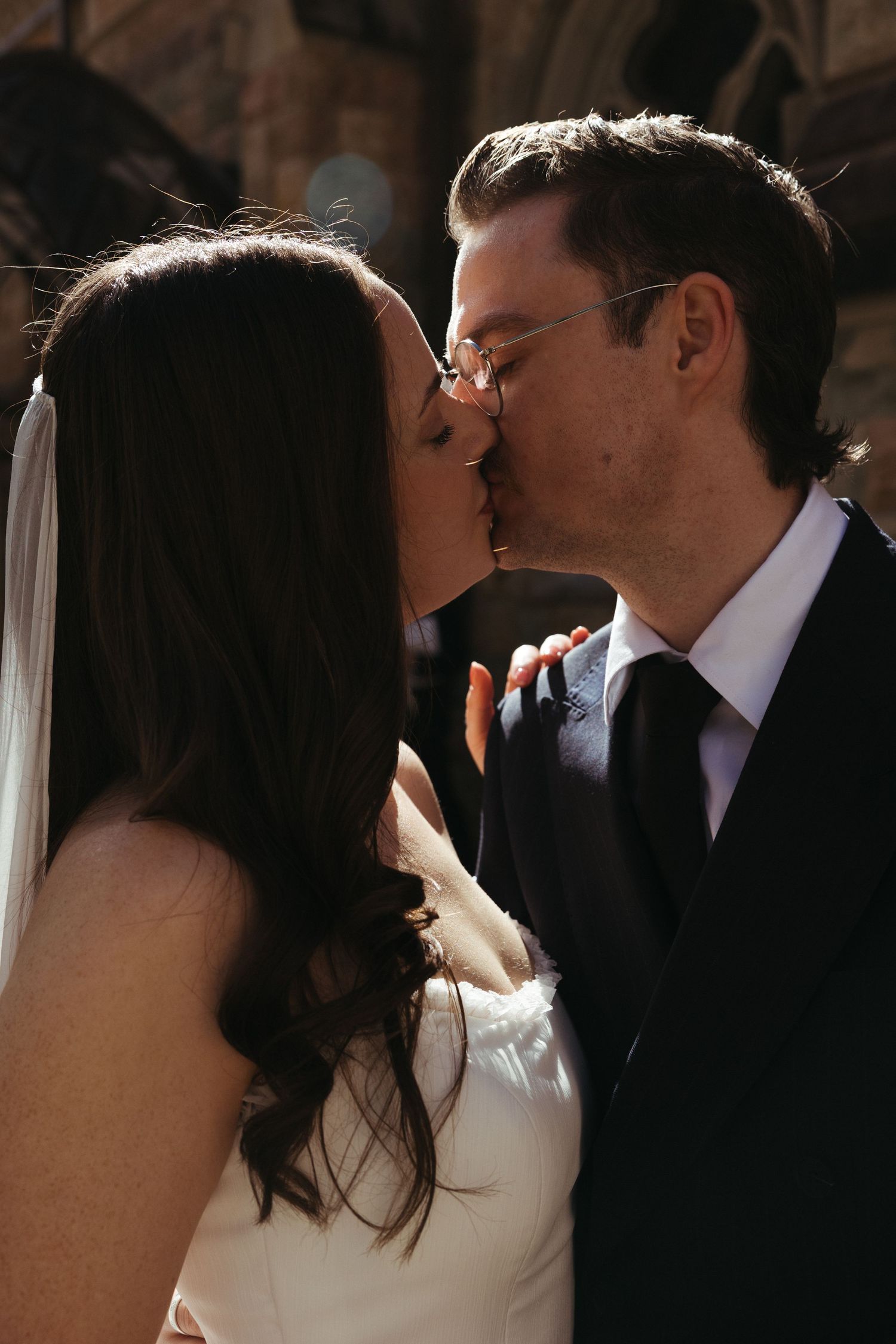 A romantic wedding kiss captured in soft sunlight between a bride in white and groom in a dark suit.