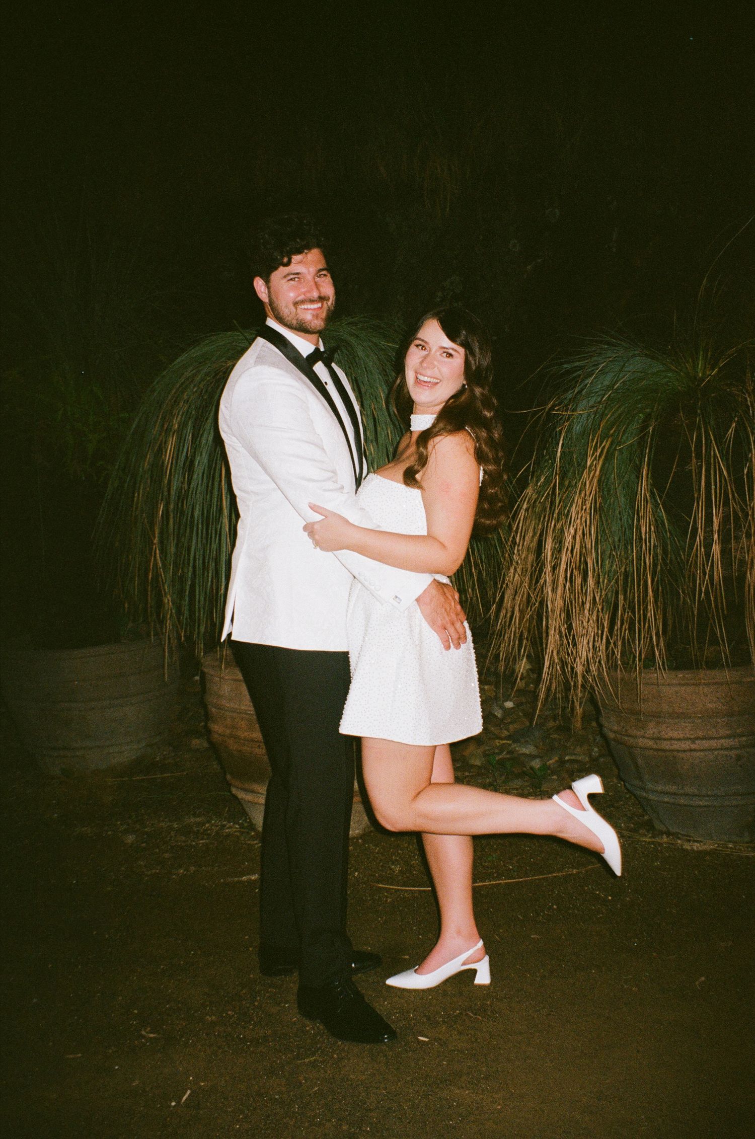 Romantic couple dancing together at night in an outdoor venue with decorative lighting and tall grasses.