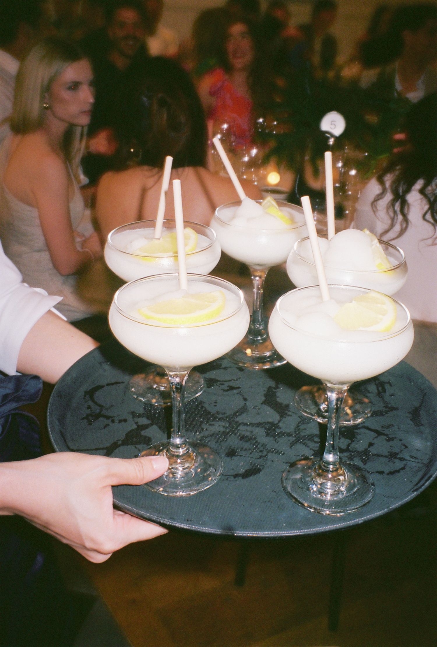 A tray of elegant cream cocktails with lemon garnishes and white straws being served.