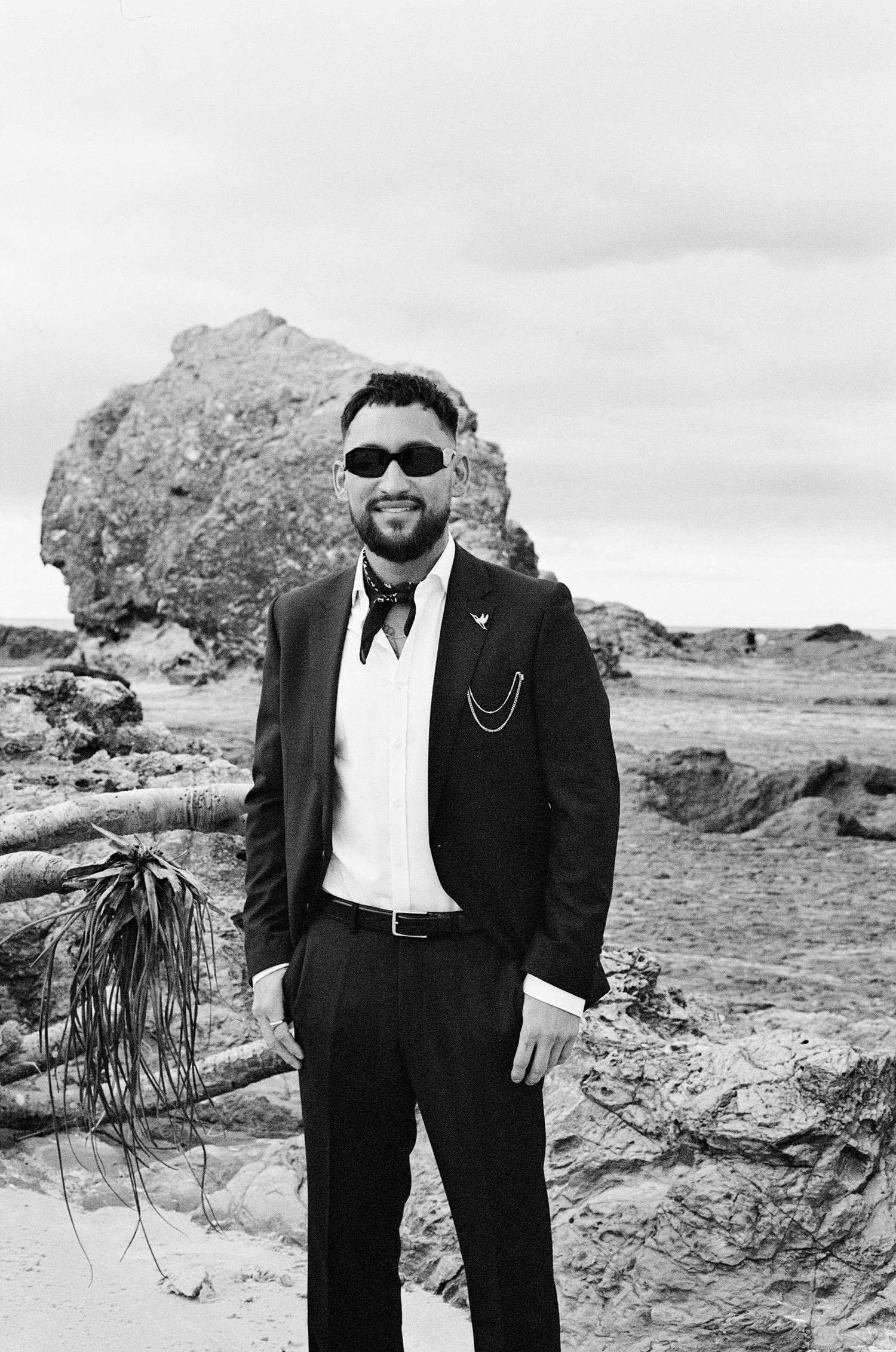 Black and white photo of someone in a formal suit standing in a desert landscape with sunglasses and a large rock formation.