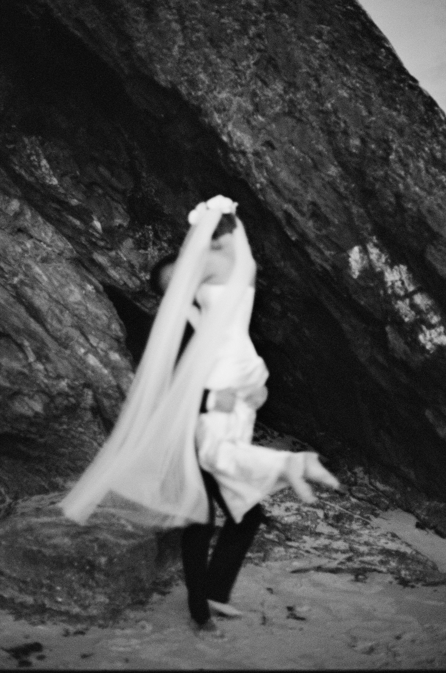 A dramatic black and white photo of someone in flowing white fabric against a rocky coastal backdrop.