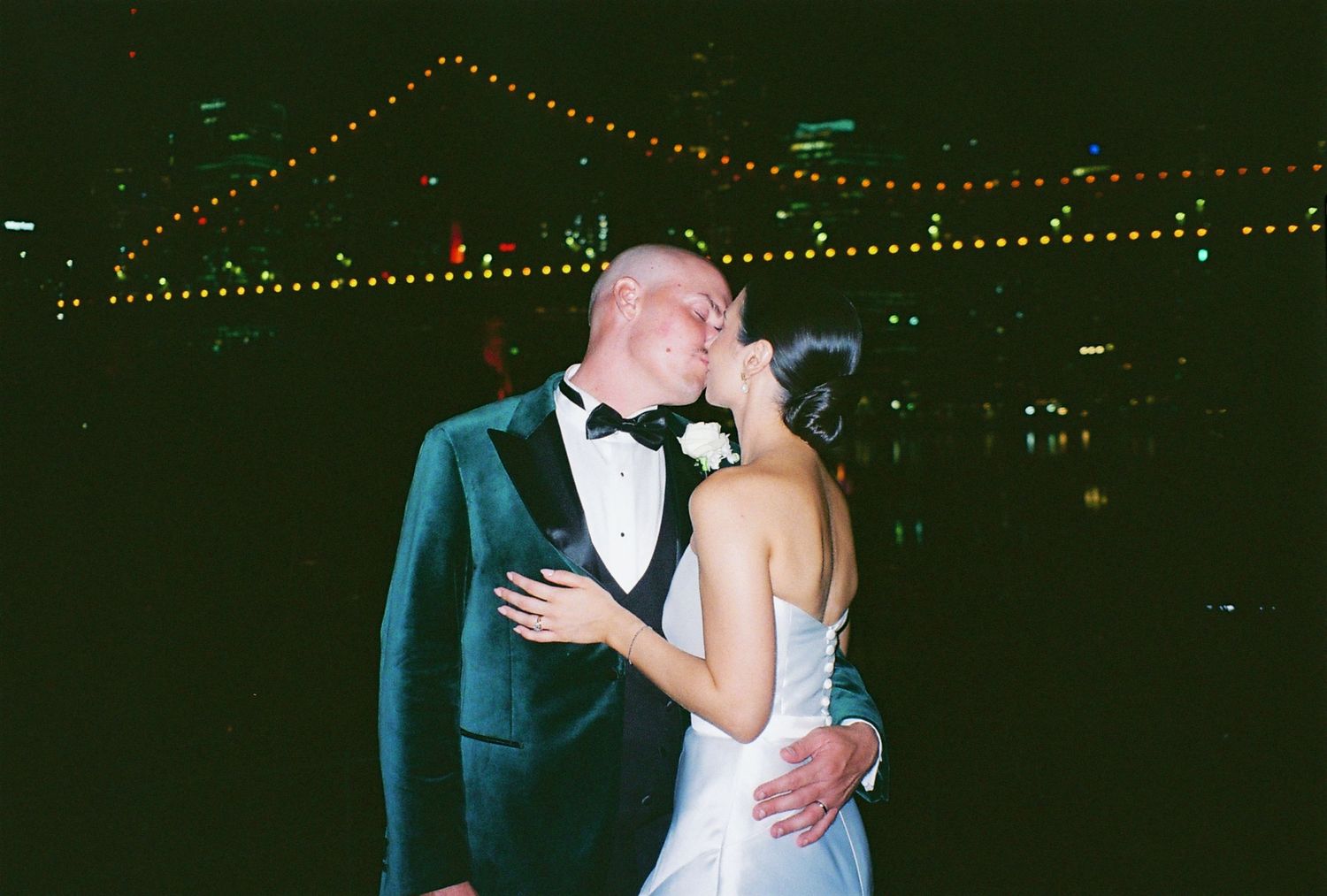 Couple embracing during romantic nighttime moment with string lights in background.