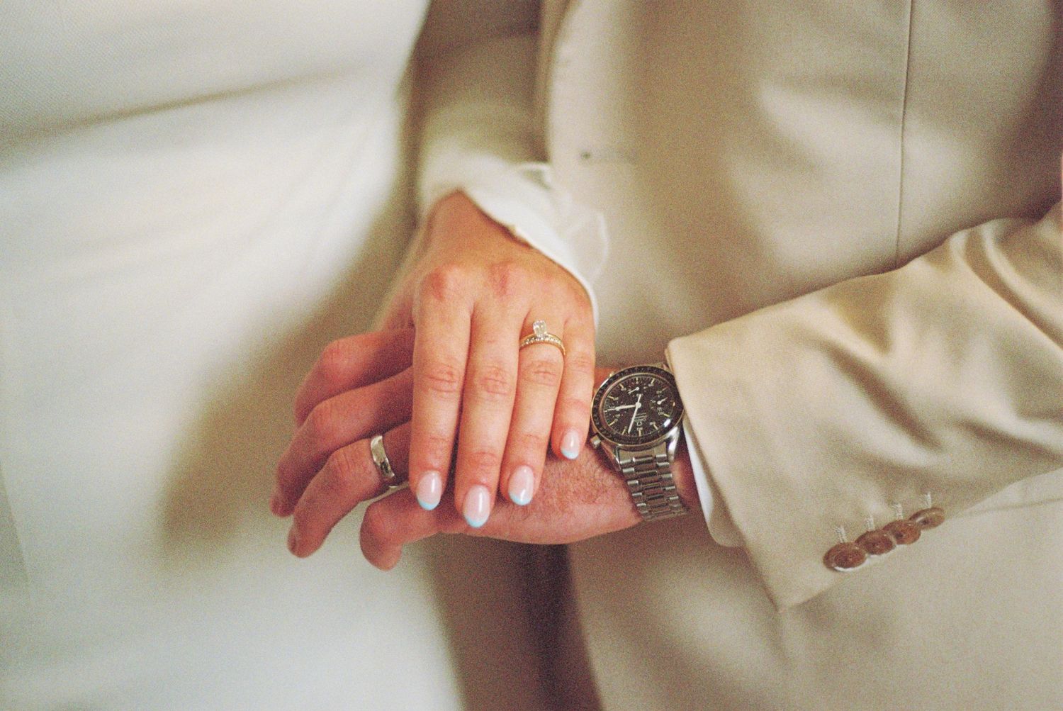 Close-up shot of hands showing wedding rings and a stylish watch against light colored formal wear.