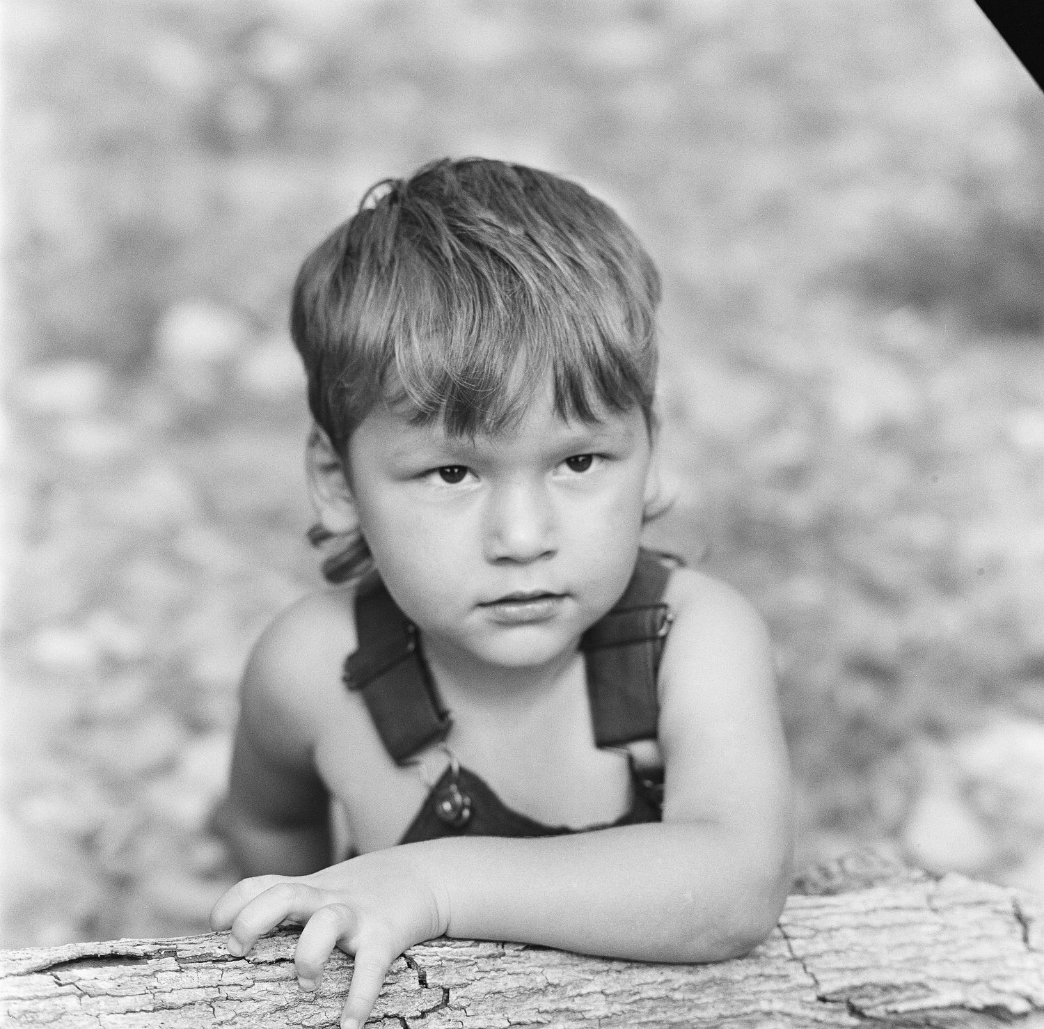 Black and white portrait of a young child in overalls leaning on a log outdoors.