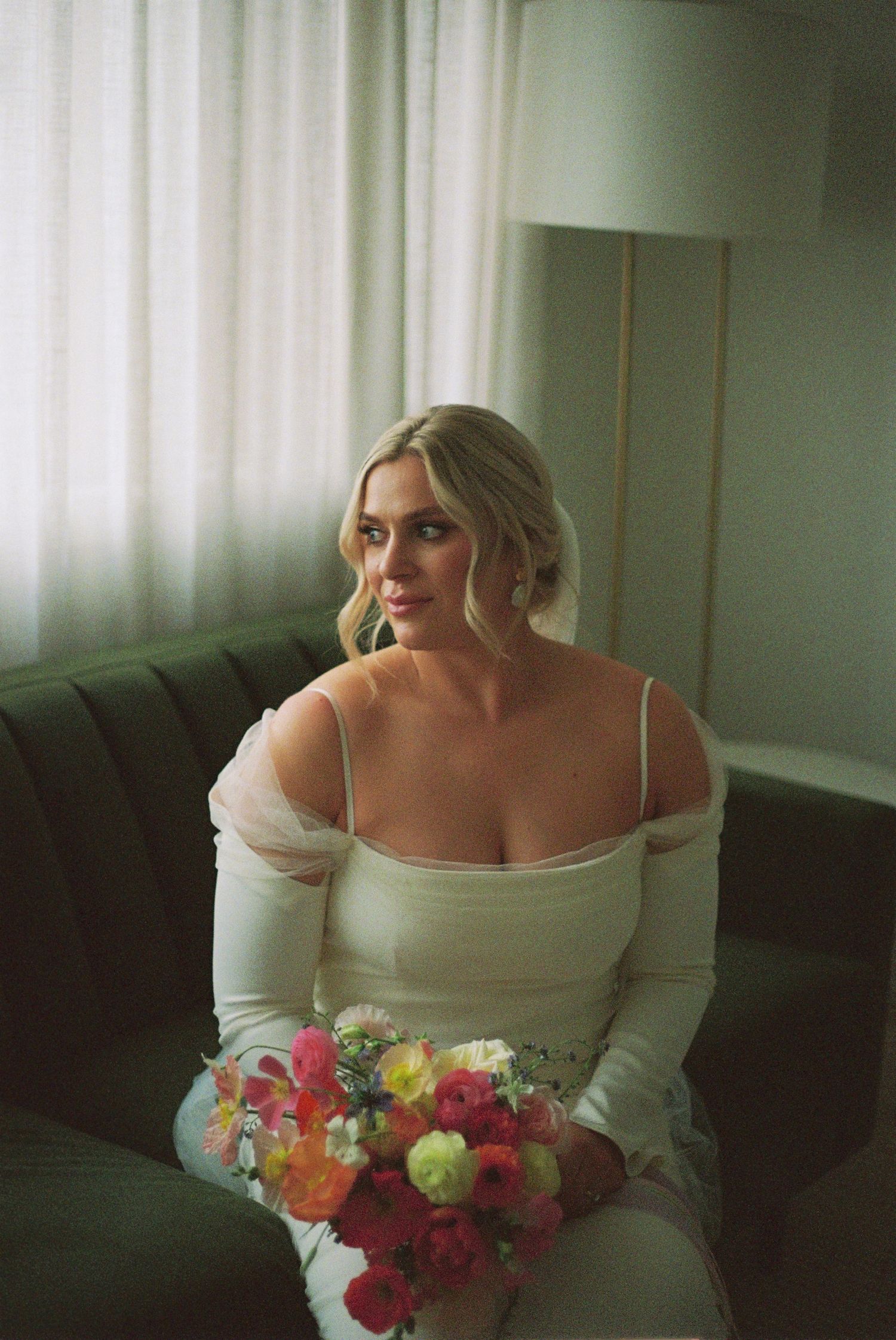 A bridal portrait on a dark couch featuring an off-shoulder dress and colorful bouquet.
