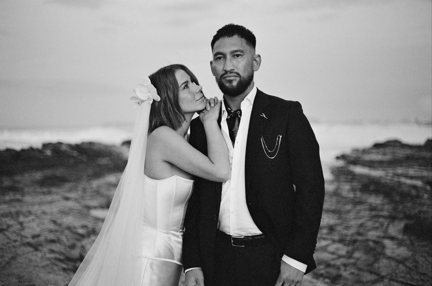 Black and white wedding portrait of a couple posing outdoors in formal attire with bride in white veil and groom in suit.