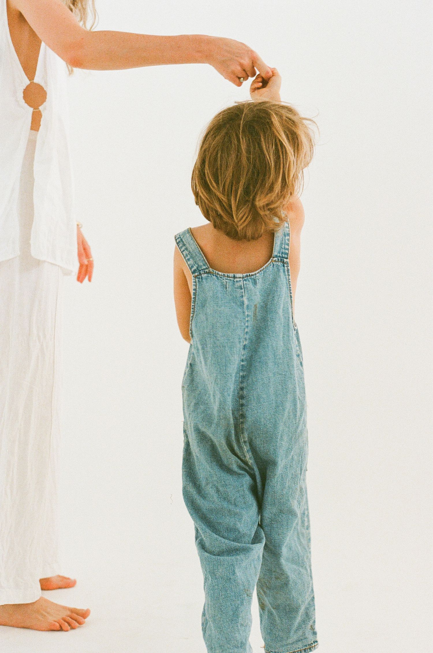 Minimalist portrait showing someone in denim overalls standing against white background.