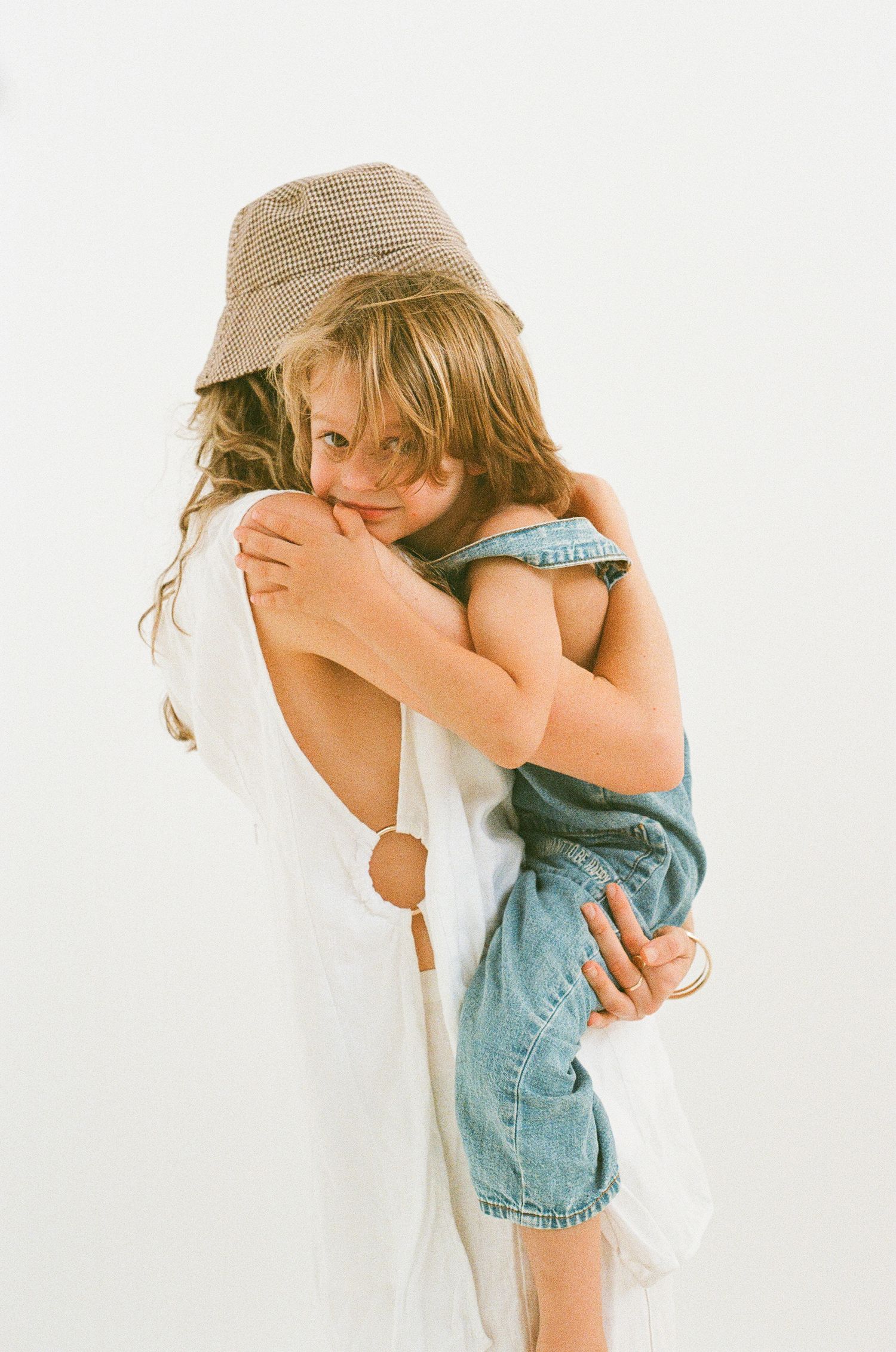 A tender embrace between parent and child wearing denim and a beige sun hat against white background.