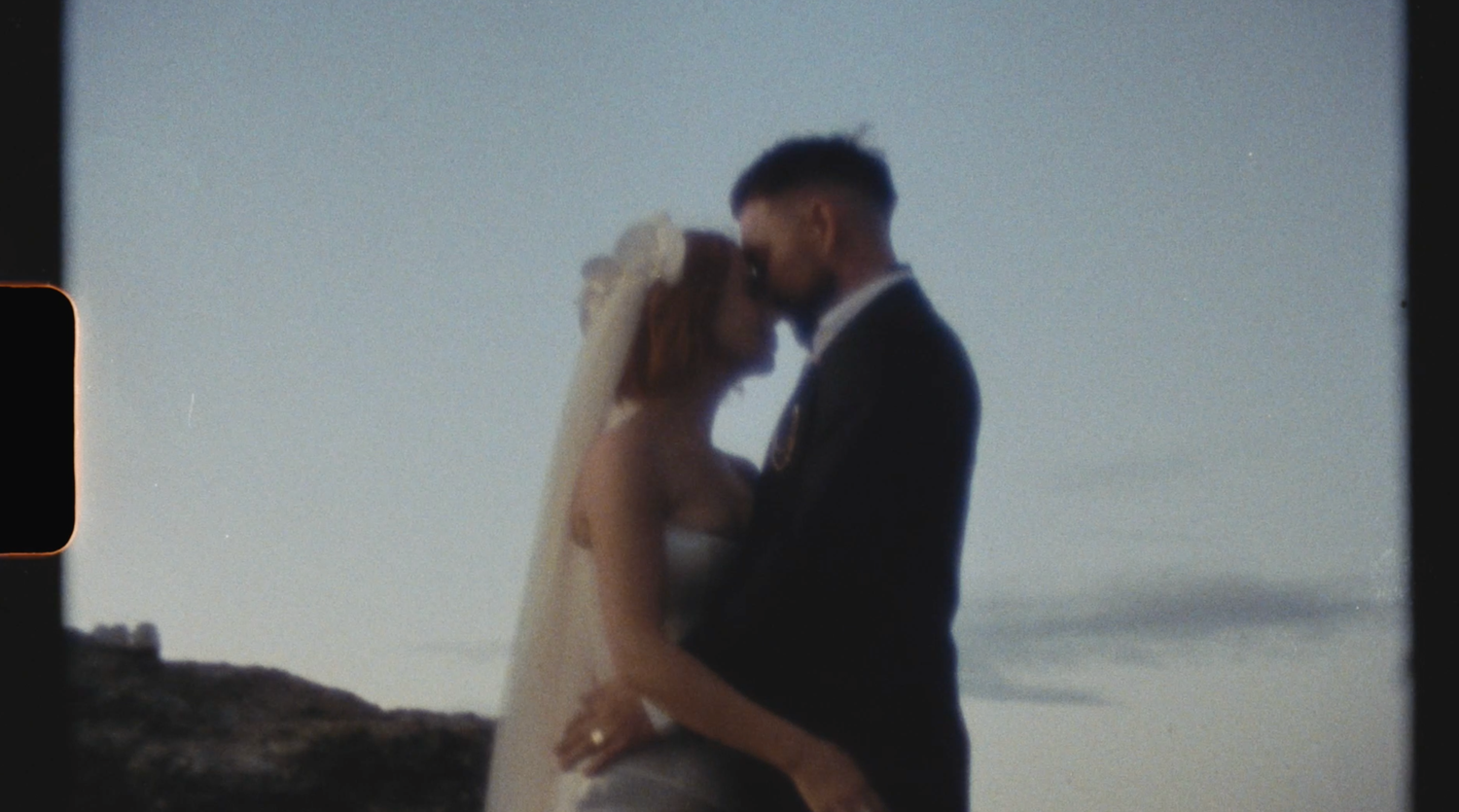 A couple shares a romantic kiss at sunset while standing on a balcony overlooking the ocean.