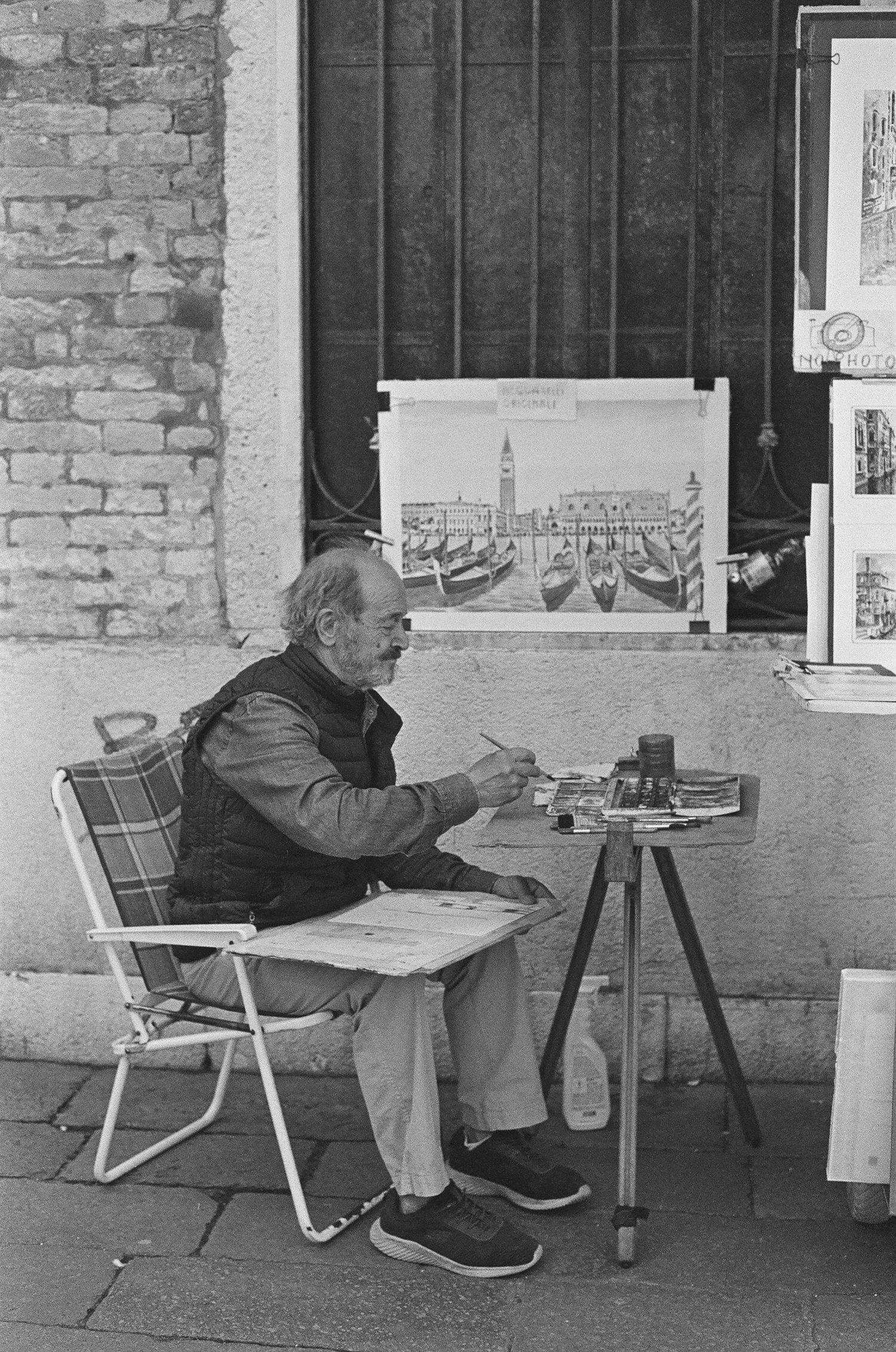 Street artist sketches Venice scene while sitting at small table with artwork display.