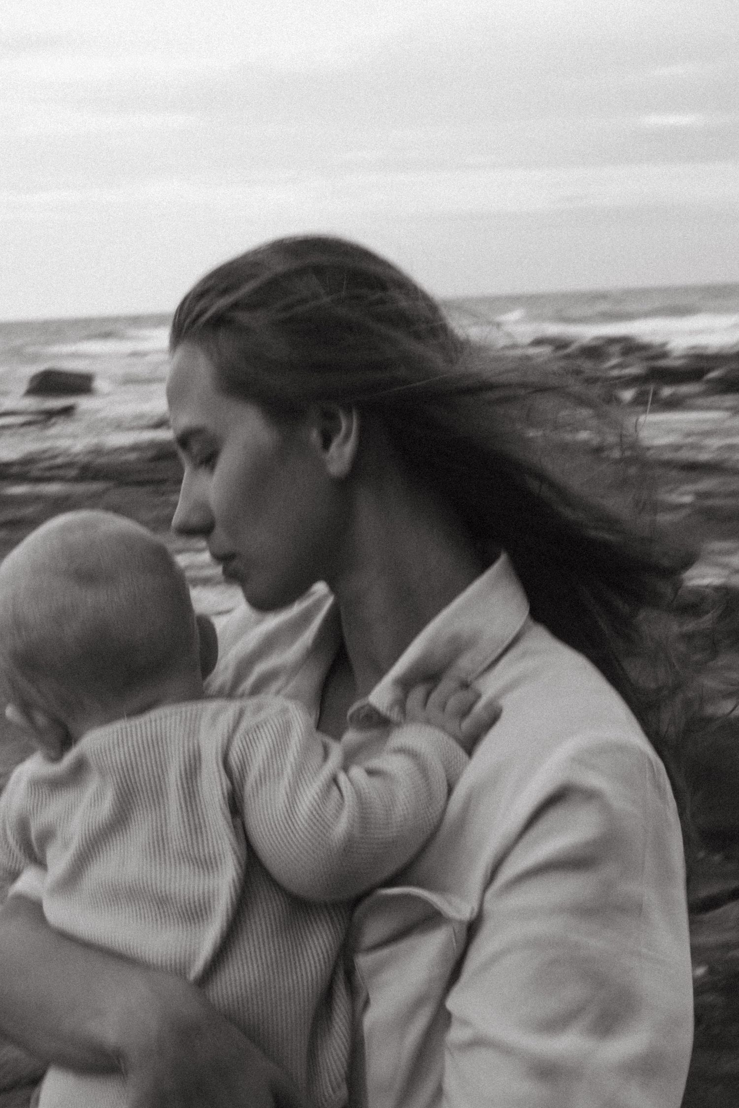 Mother in light coat holding baby against dramatic oceanside backdrop in black and white.
