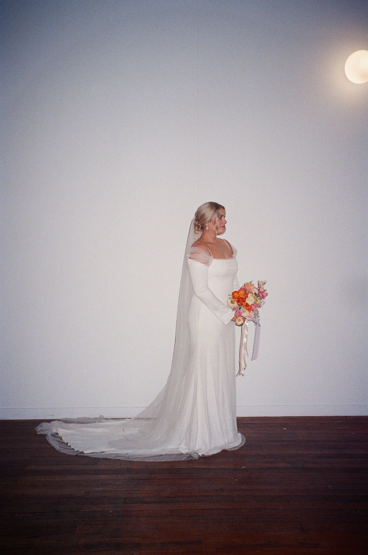 A bride in an elegant white off-shoulder wedding gown holding a vibrant orange bouquet poses against a light background.