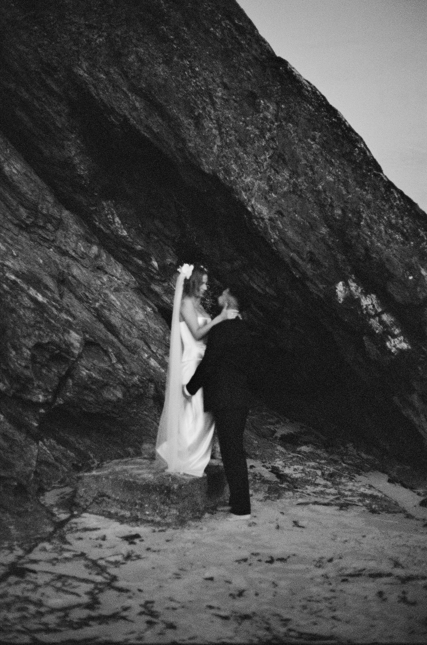 Black and white series showing a couple walking and dancing in the shadows of a dramatic rocky cave formation by the beach.