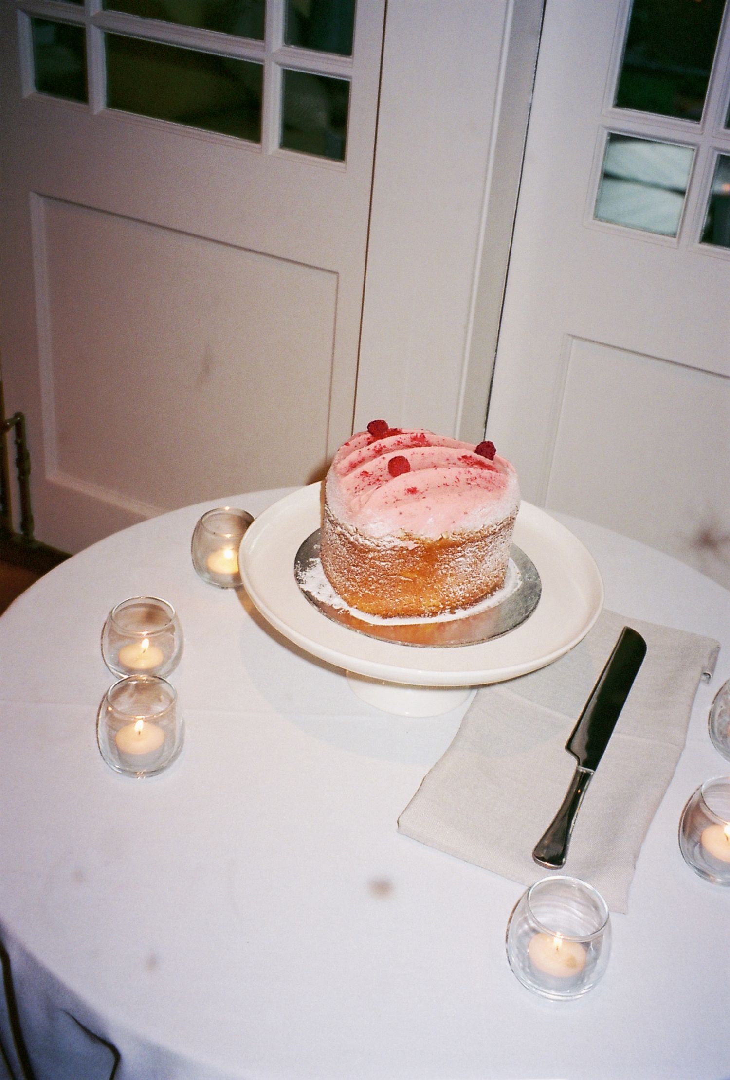 A pink-frosted cake sits on a white table surrounded by small candlelit votives.