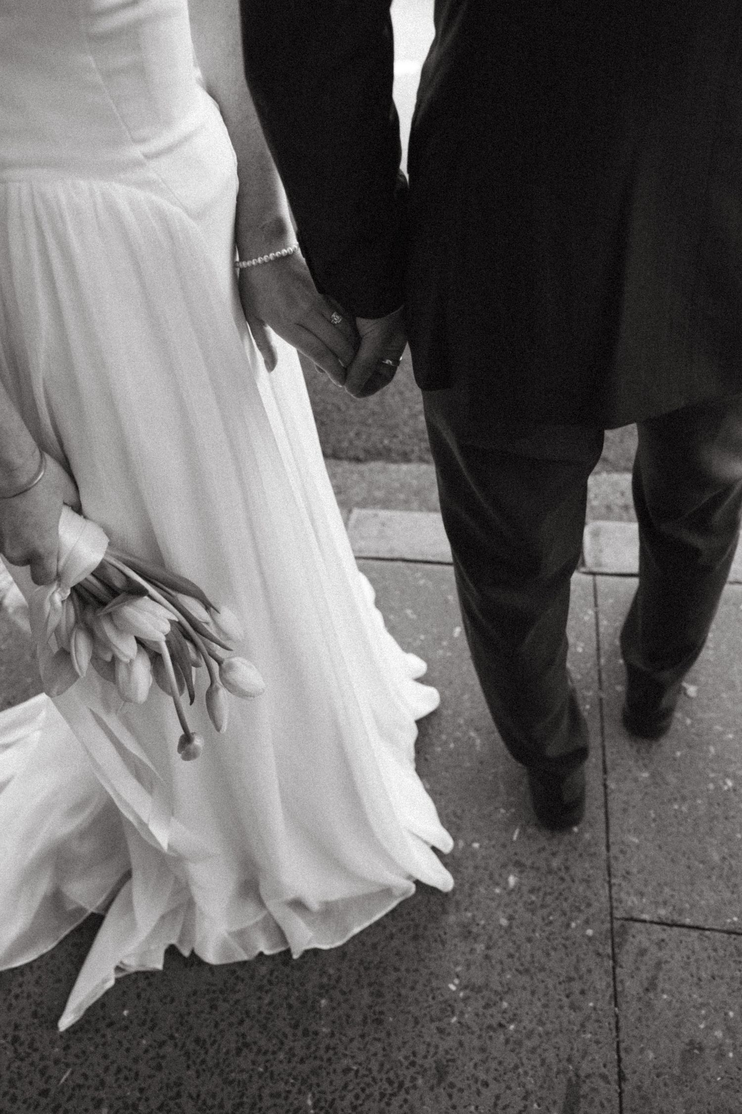 Black and white wedding photo showing a couple holding hands, with bride in flowing white dress holding flowers.