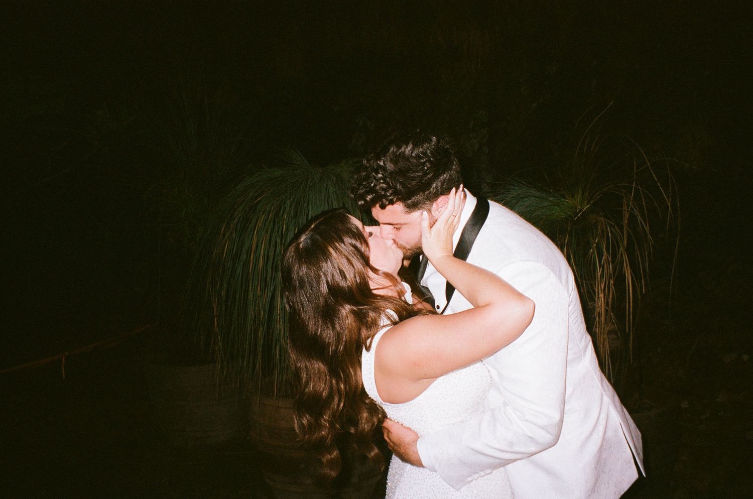 A romantic couple in white clothing embrace and kiss outdoors at night.