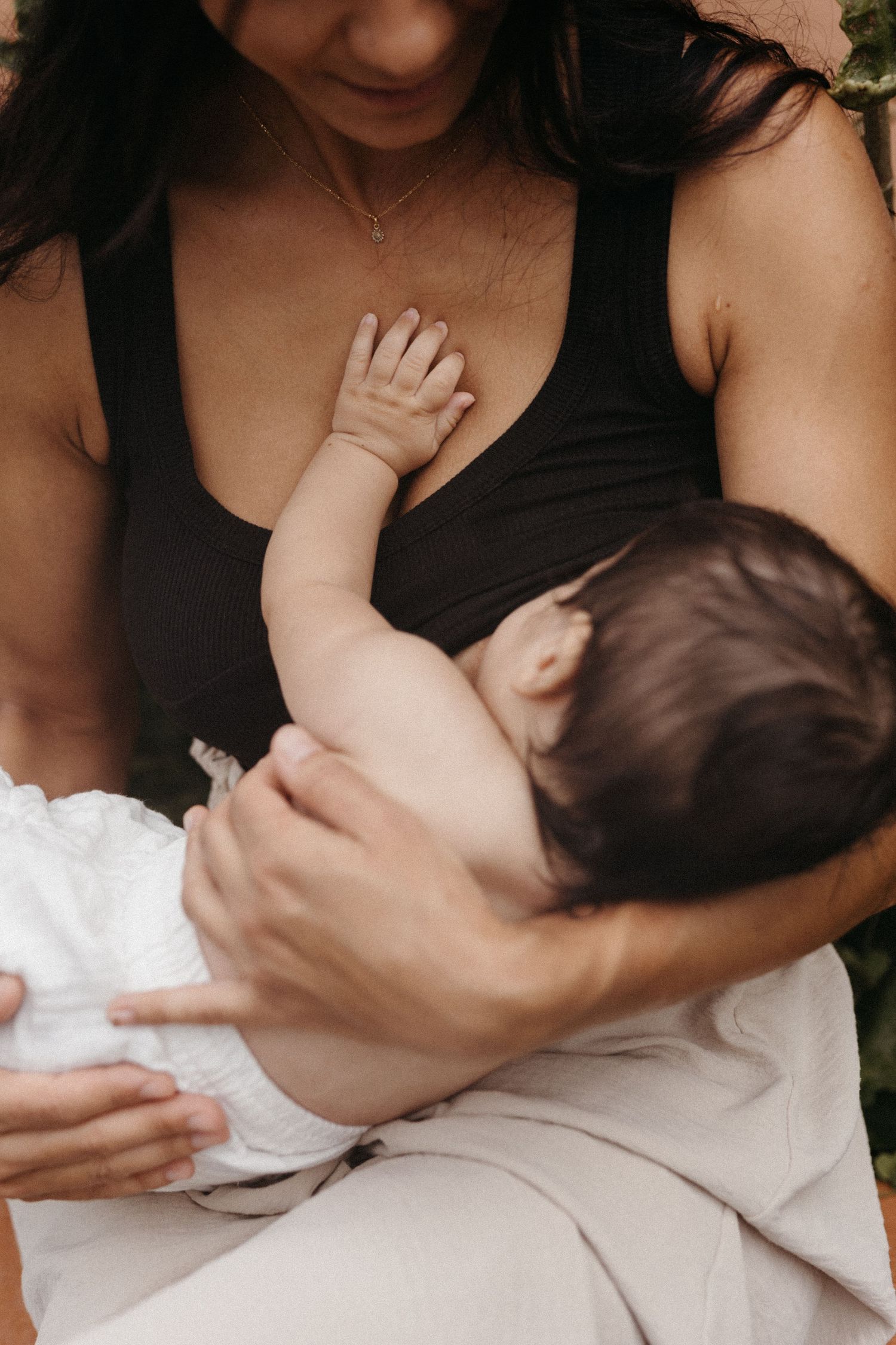 Intimate close-up of parent holding nursing baby in black tank top against natural backdrop.