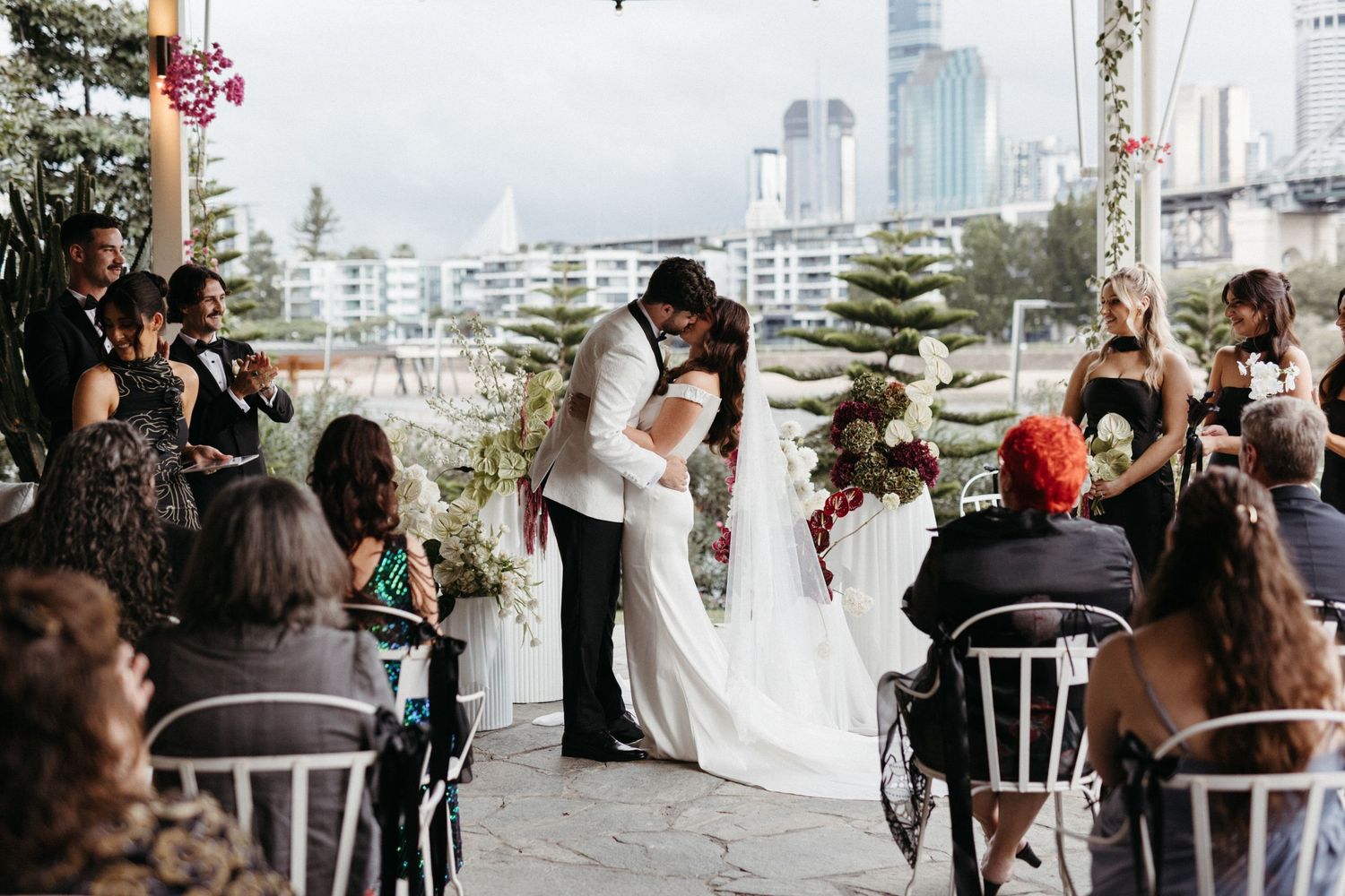 A couple shares their first kiss at an intimate outdoor waterfront wedding ceremony while guests look on from decorated chairs.