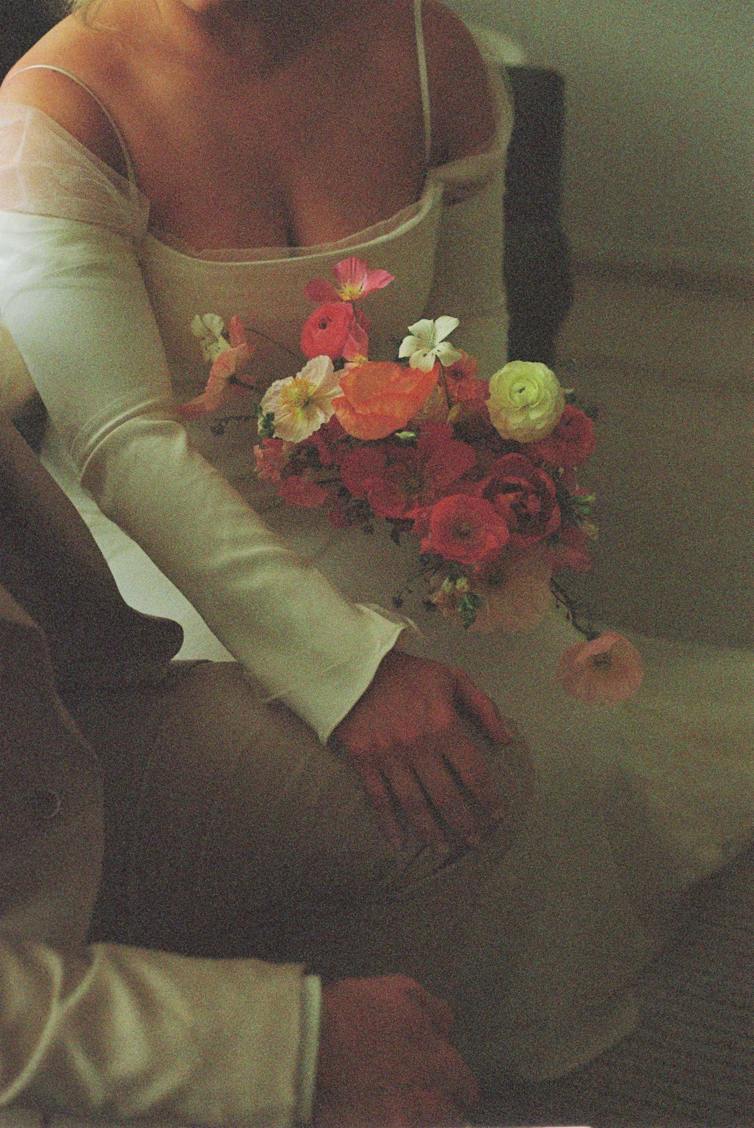 Artistic shot of a floral bouquet with red and coral blooms held by someone in cream-colored sleeves.