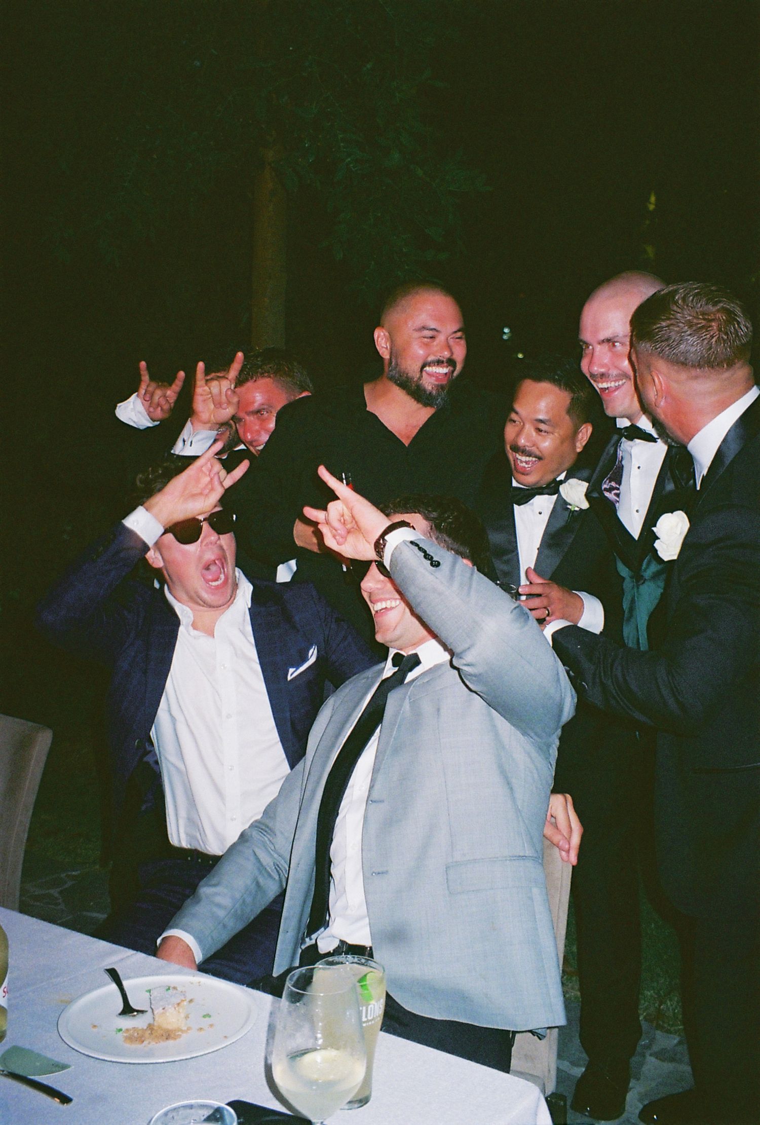 A lively group in formal attire making playful gestures while celebrating at a dinner table.