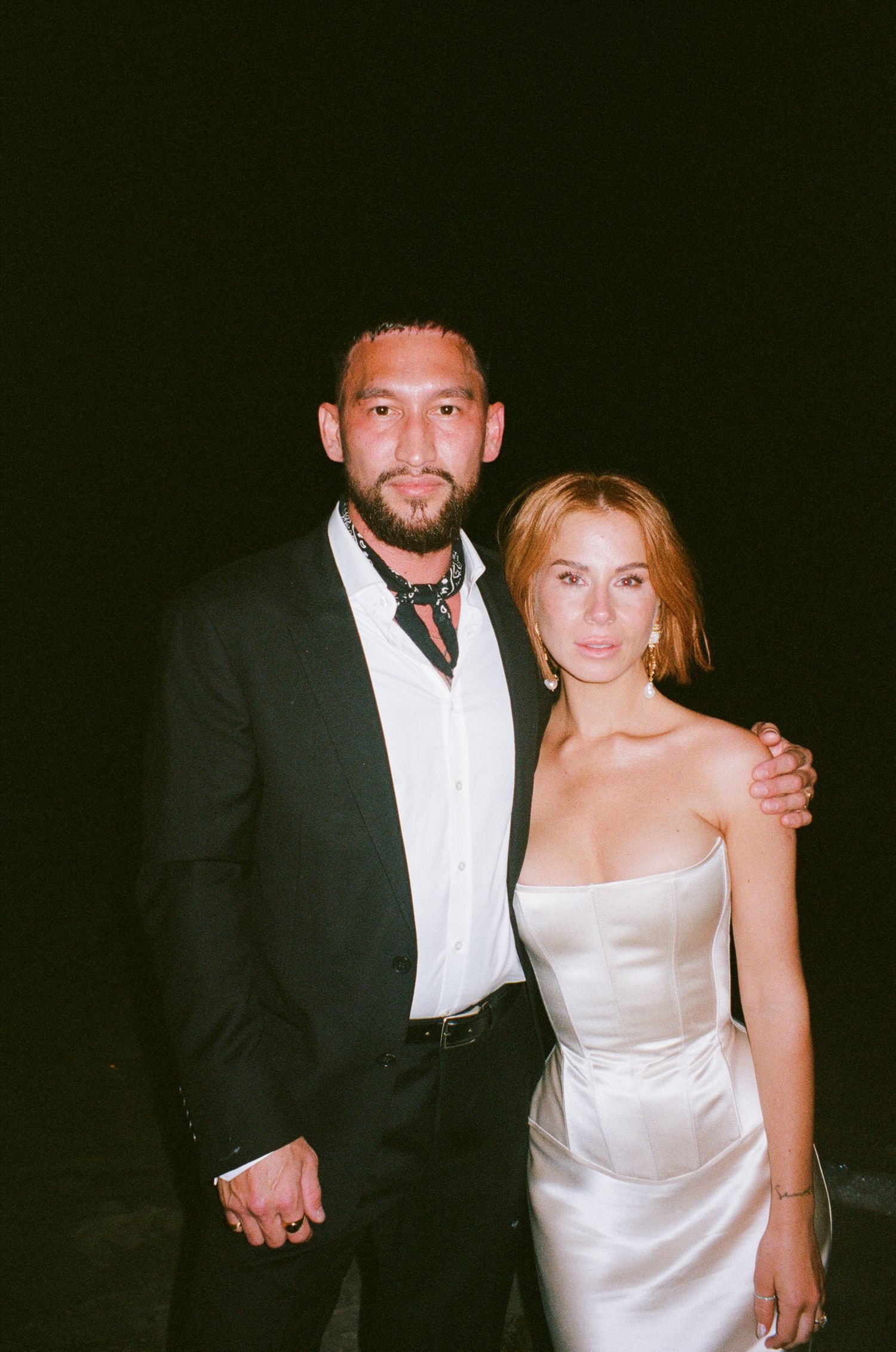 Two people pose together at a formal event with dim lighting, one wearing a black tuxedo and the other in a white dress.