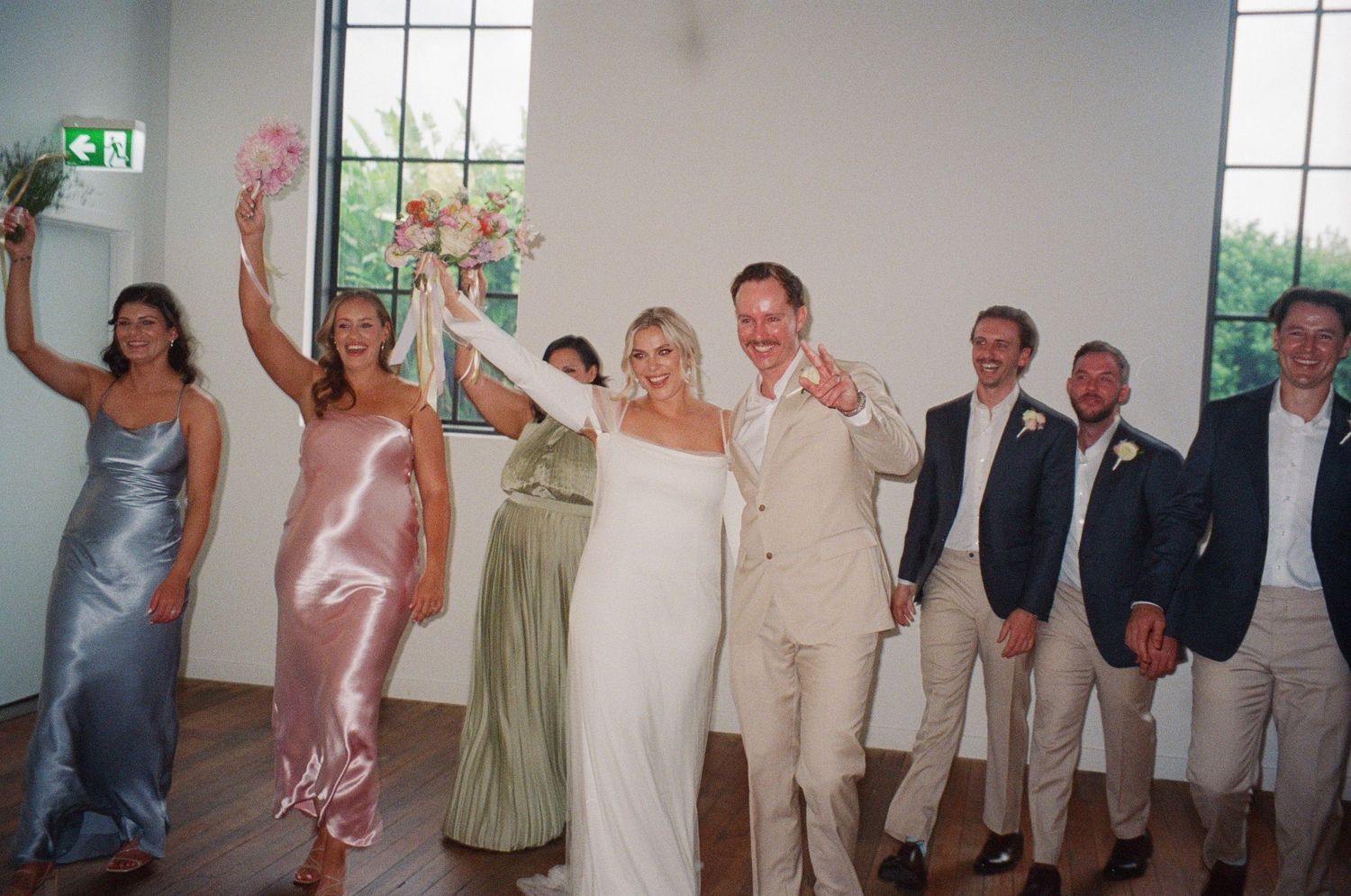 Wedding party members in coordinated attire celebrating with raised arms in a bright venue.