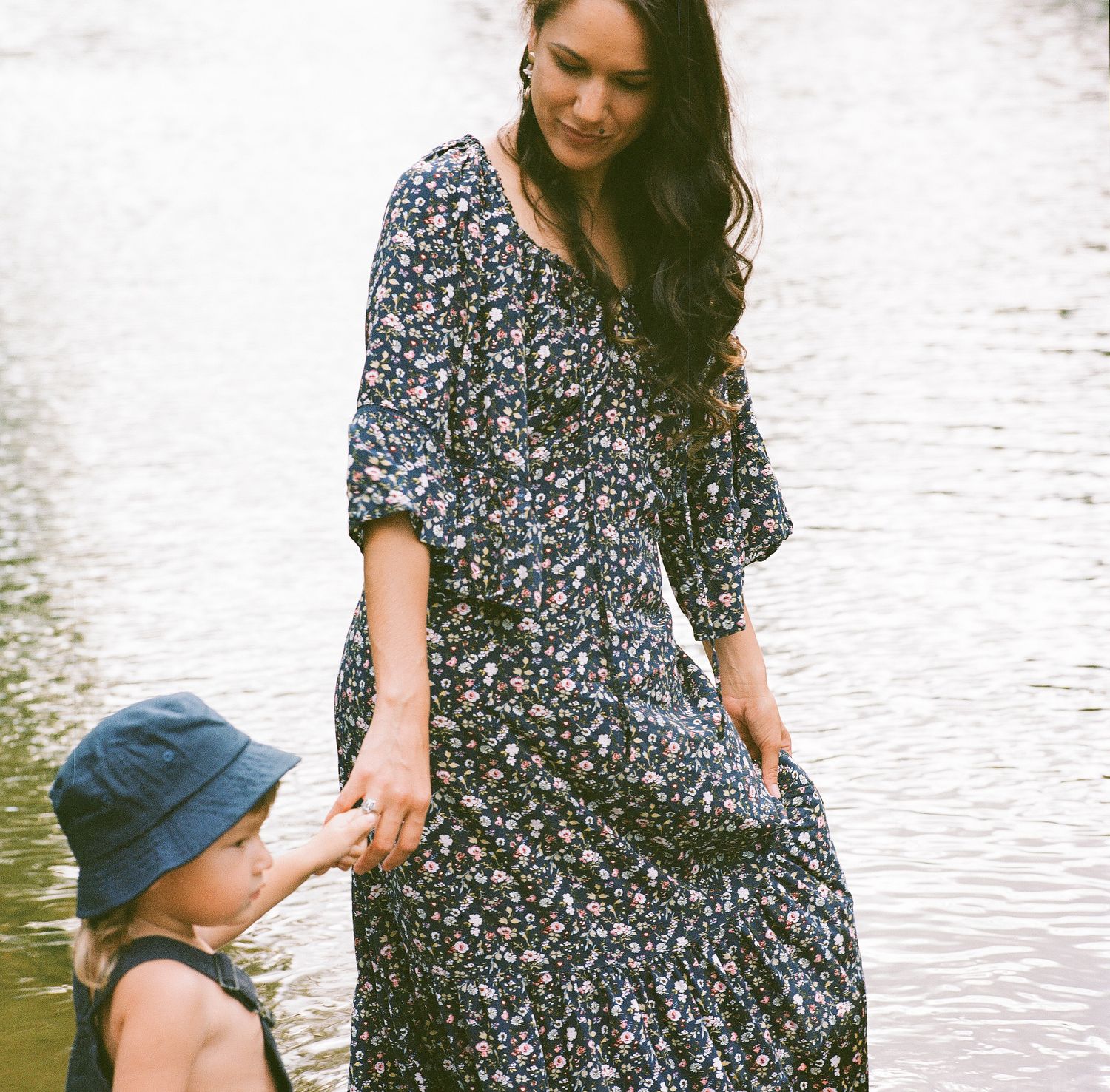 Mother in floral dress holding child's hand by tranquil waterside.