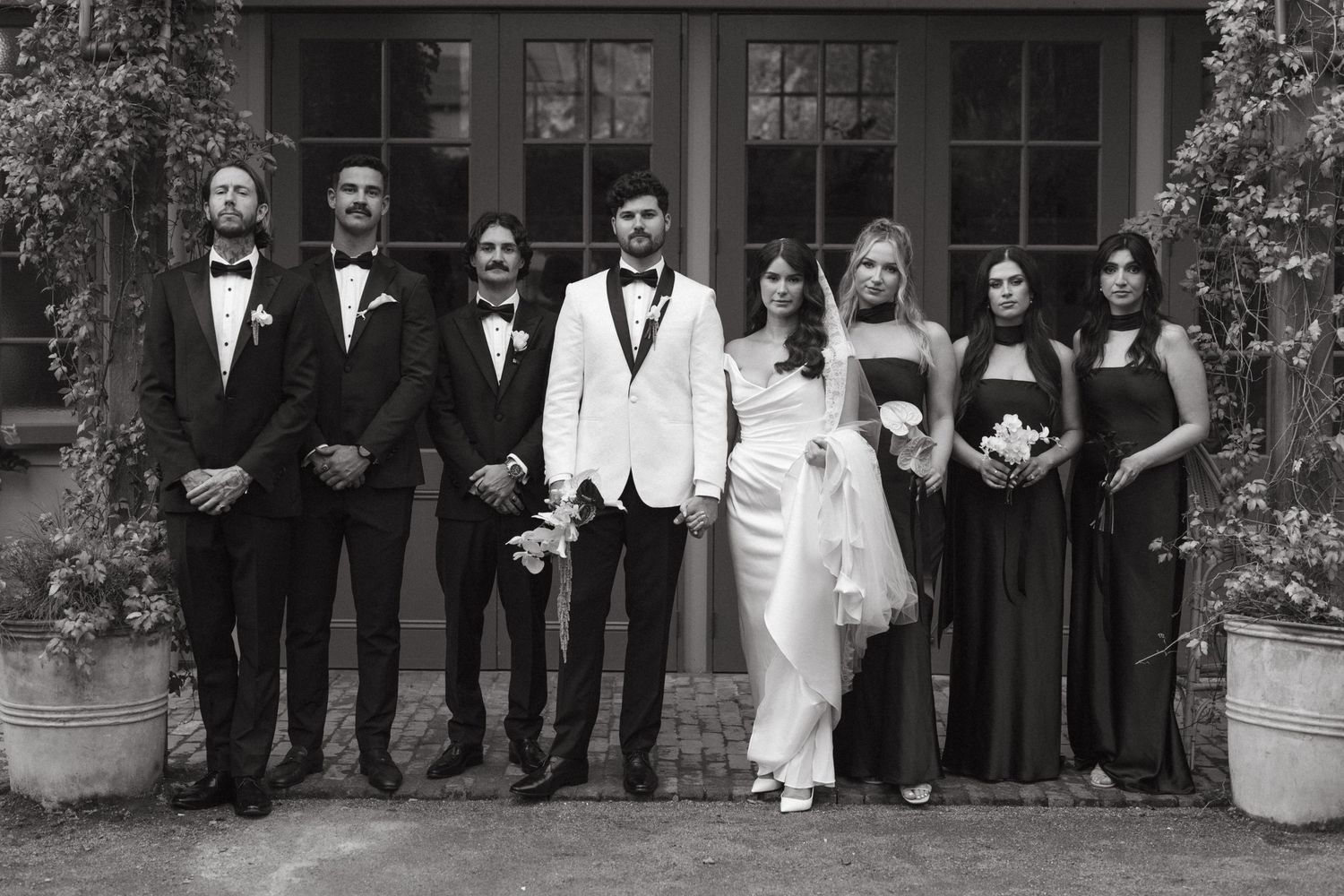 A wedding party poses in formal attire outside an elegant stone building with vintage windows and foliage.