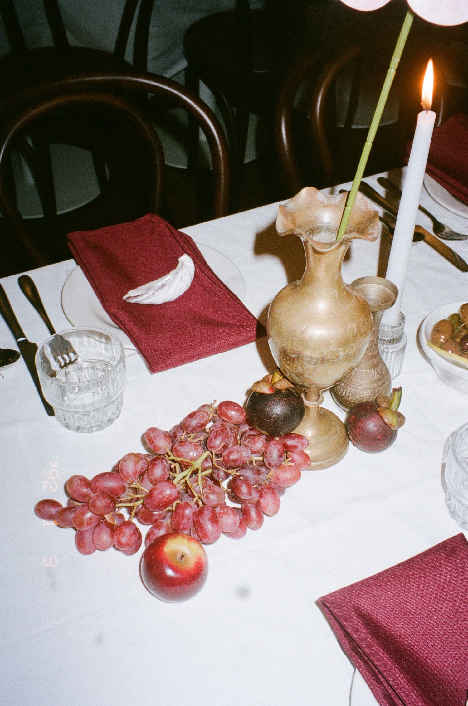 Table setting with red grapes, apples and decorative candle holders on white tablecloth with burgundy napkins.