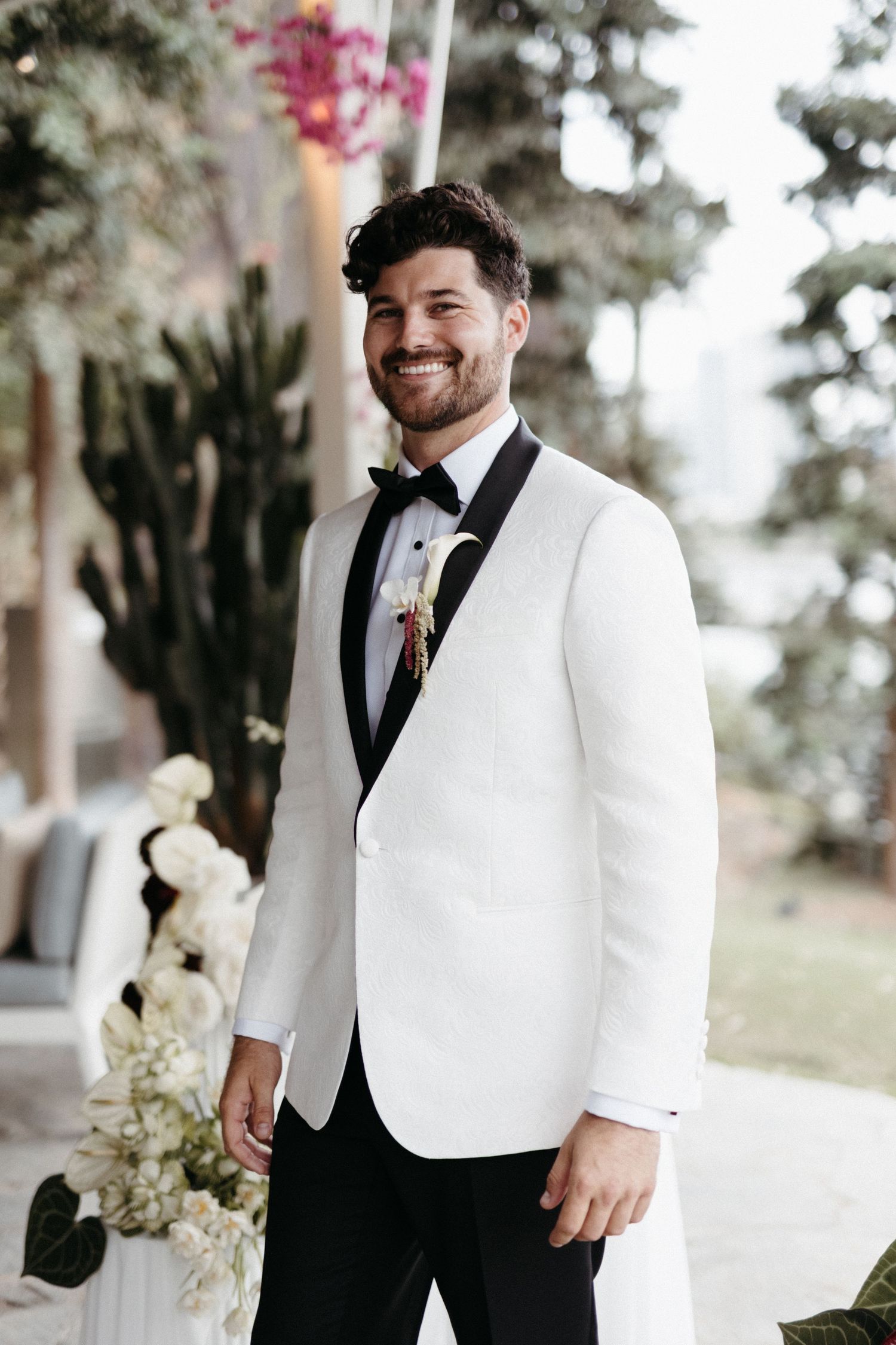 Groom wearing crisp white tuxedo jacket with black bow tie and lapel at outdoor wedding venue.