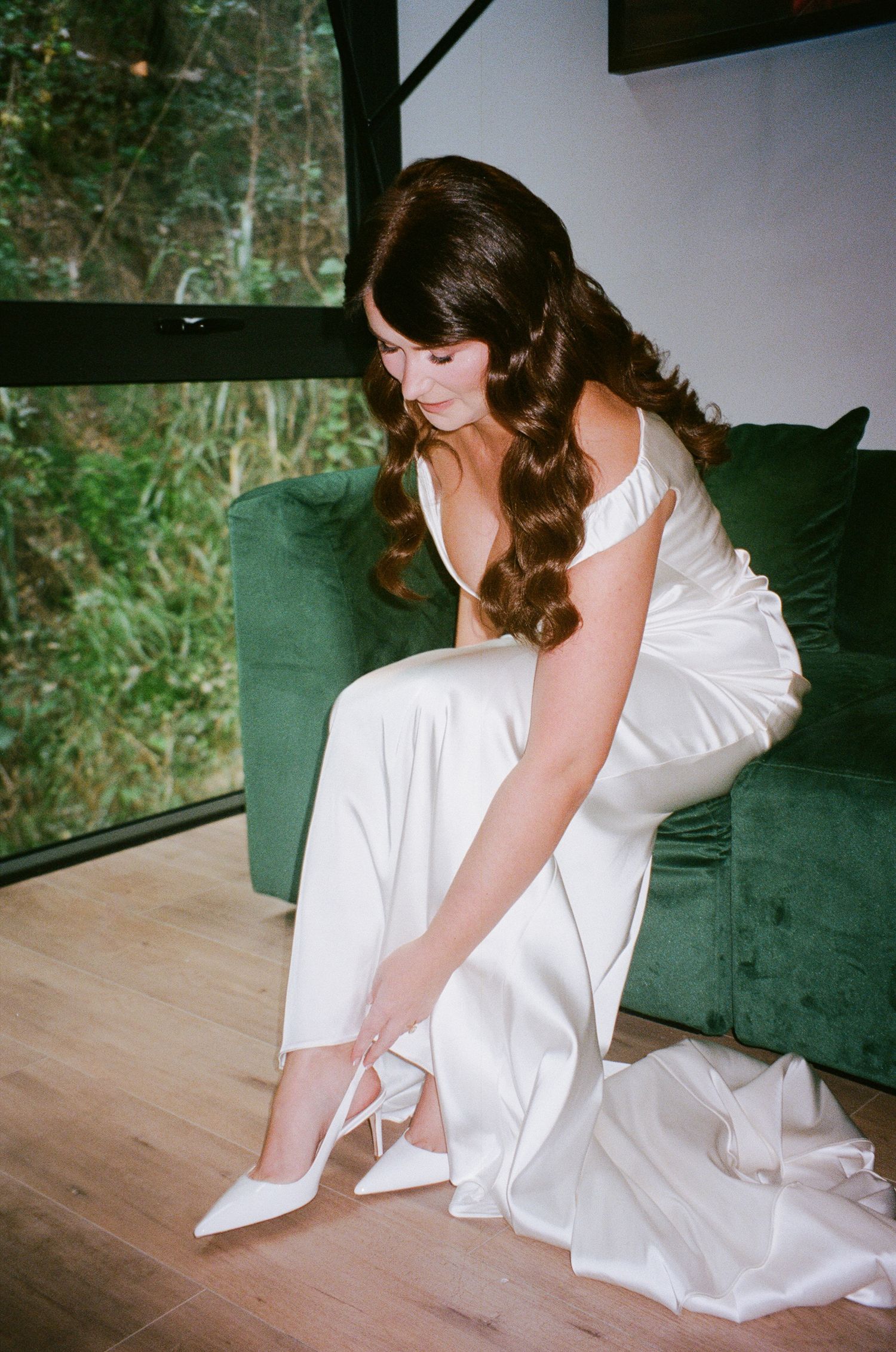 A bride in a white wedding dress adjusts her shoe on a wooden floor during wedding preparations.