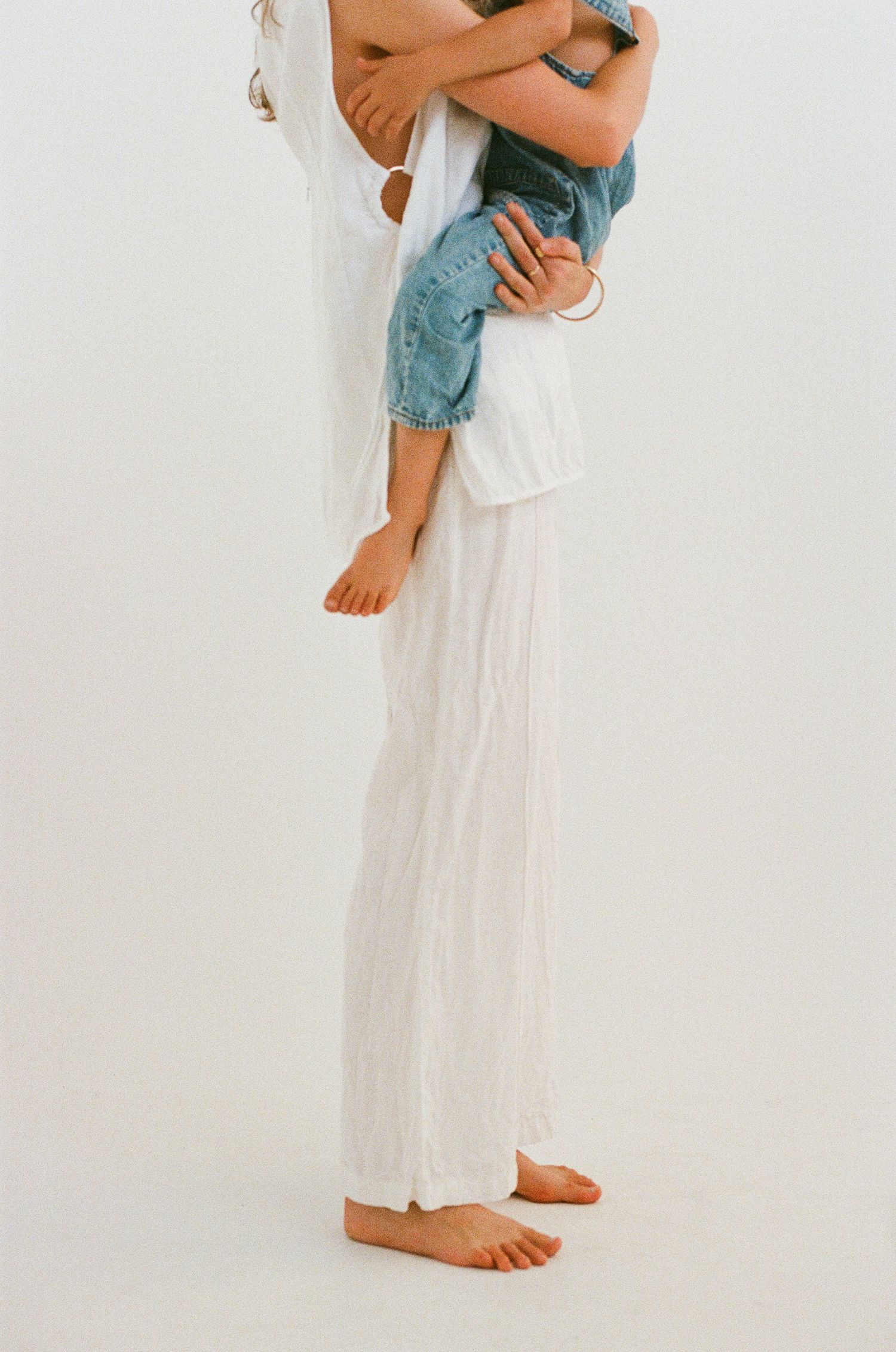 Side view of someone wearing flowing white pants and holding a child in blue overalls.