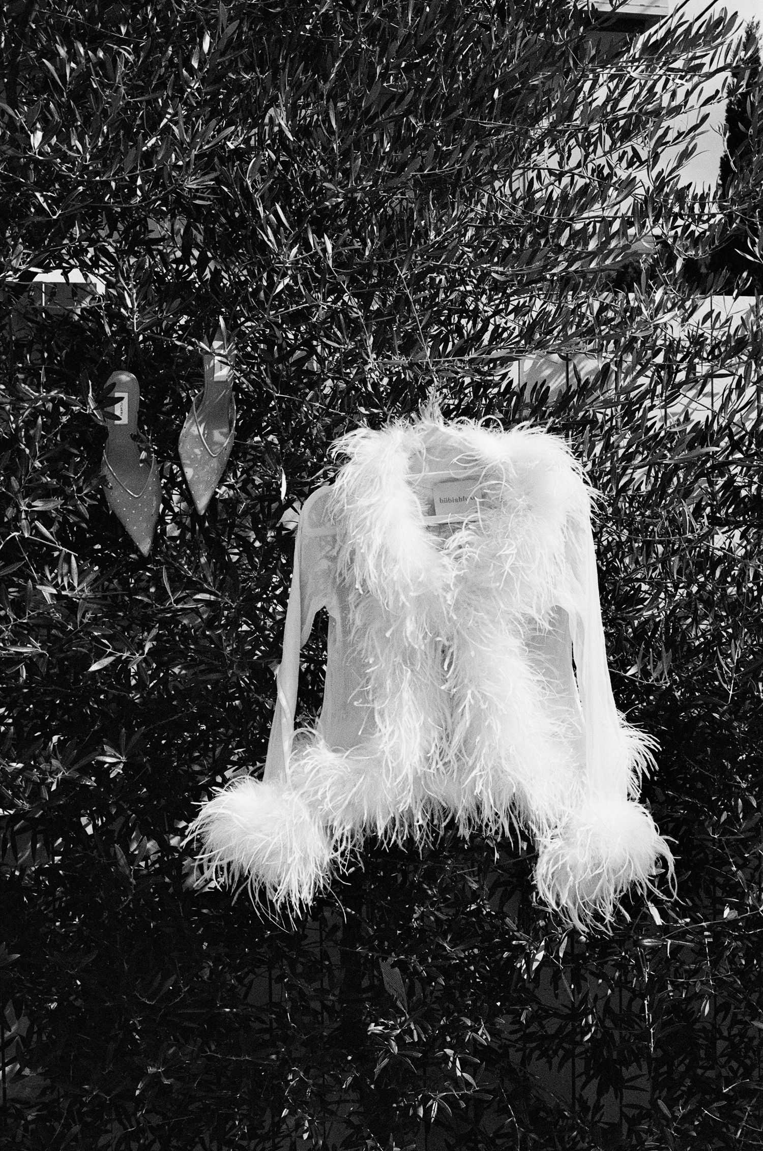 A white feathered cropped jacket with long fluffy sleeves hangs against a dark foliage background in black and white.