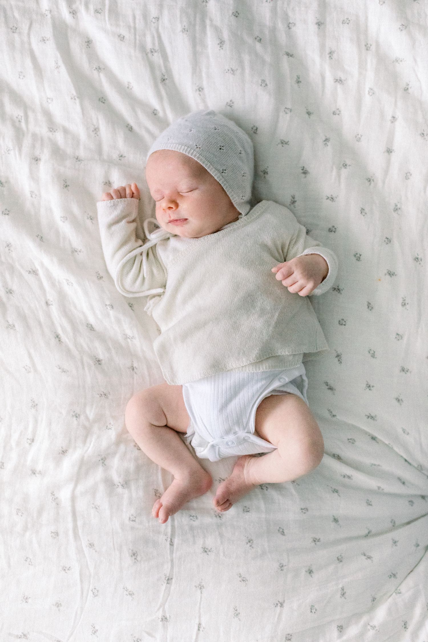 A newborn baby is peacefully sleeping on a bed in a white cashmere cardigan and a matching bonnet.