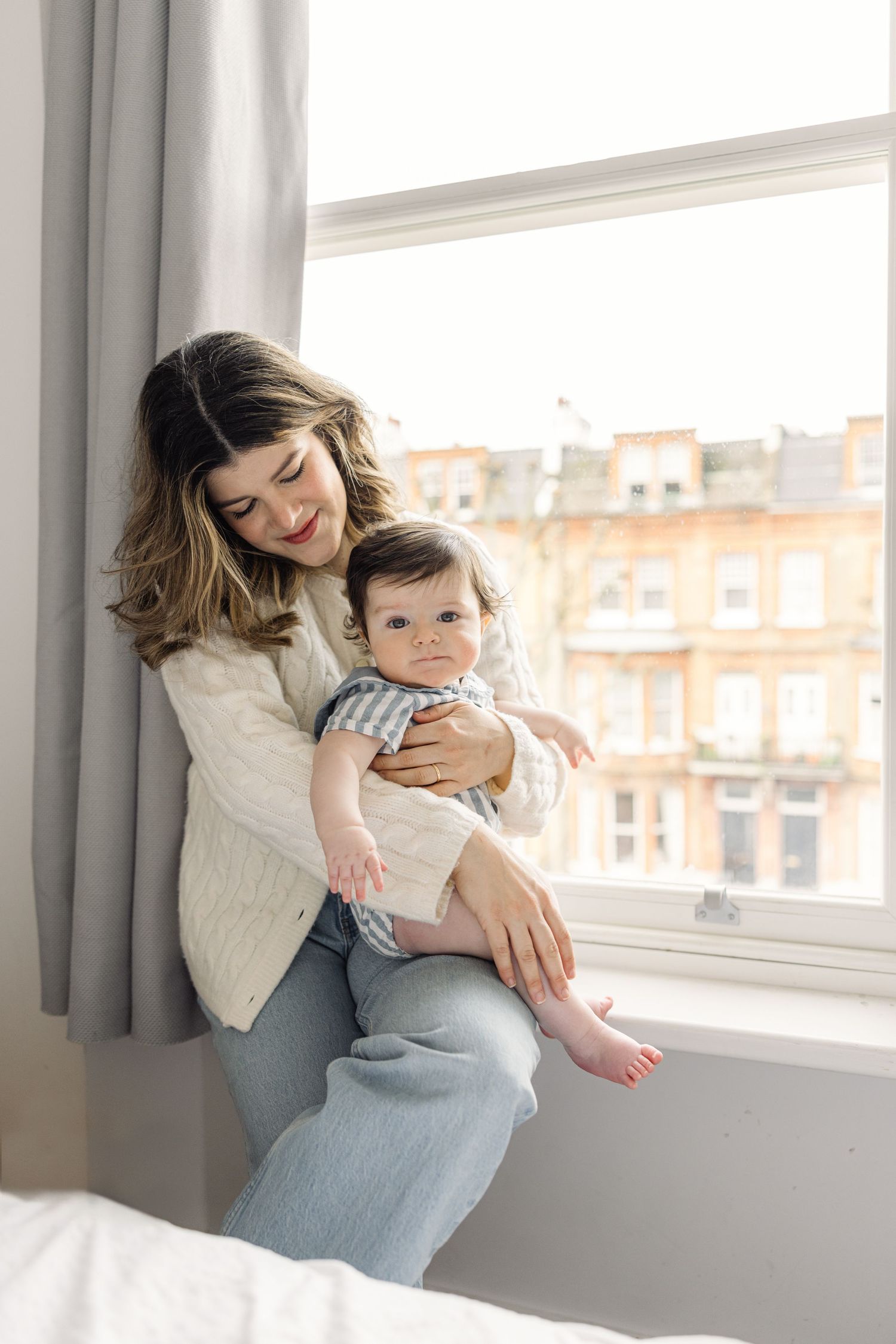 natural light baby photography in London - mother cuddling infant by window in cosy home setting