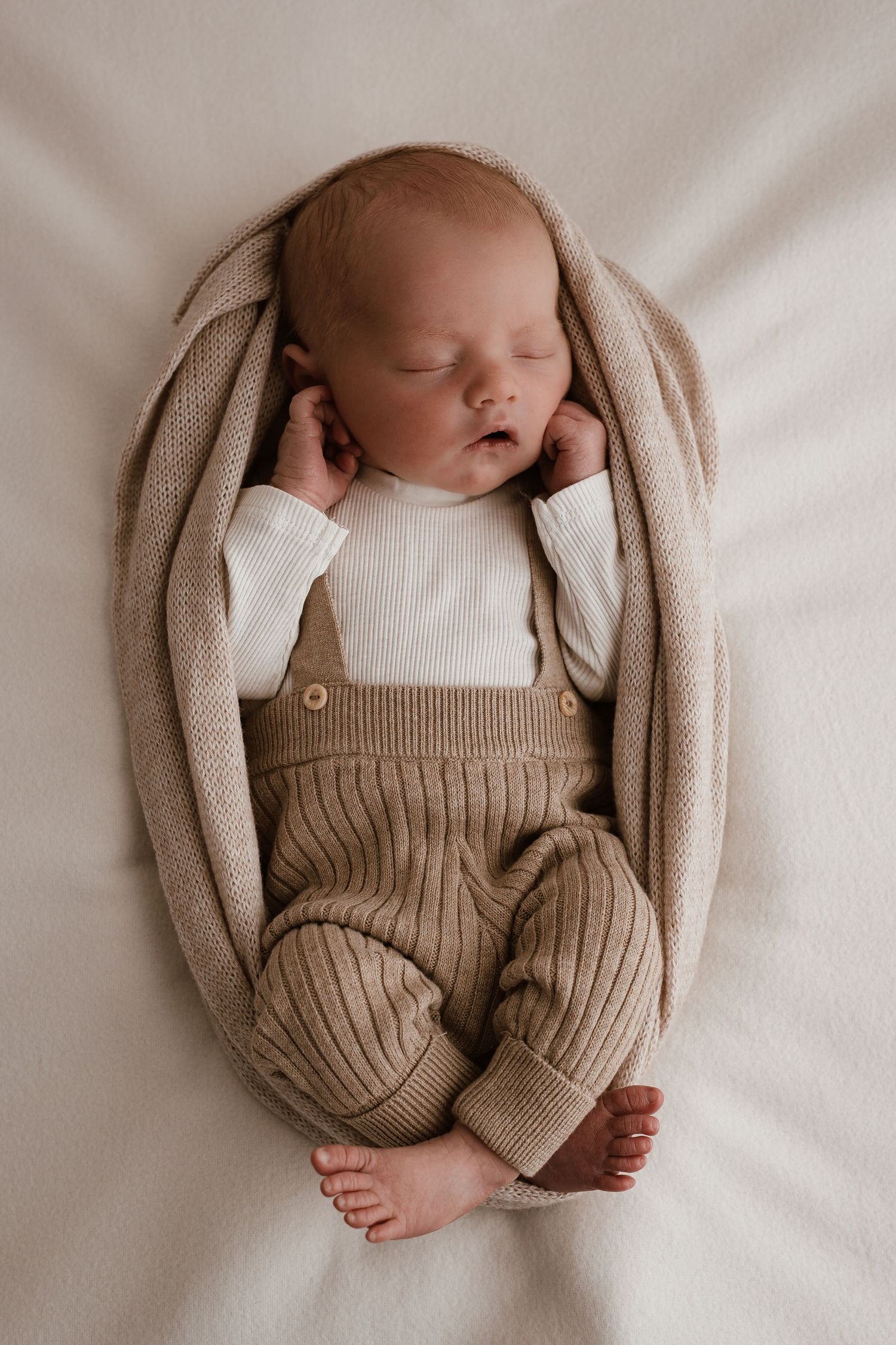 Sleeping newborn baby in beige knit overalls and white onesie, wrapped in a soft tan blanket on white background.