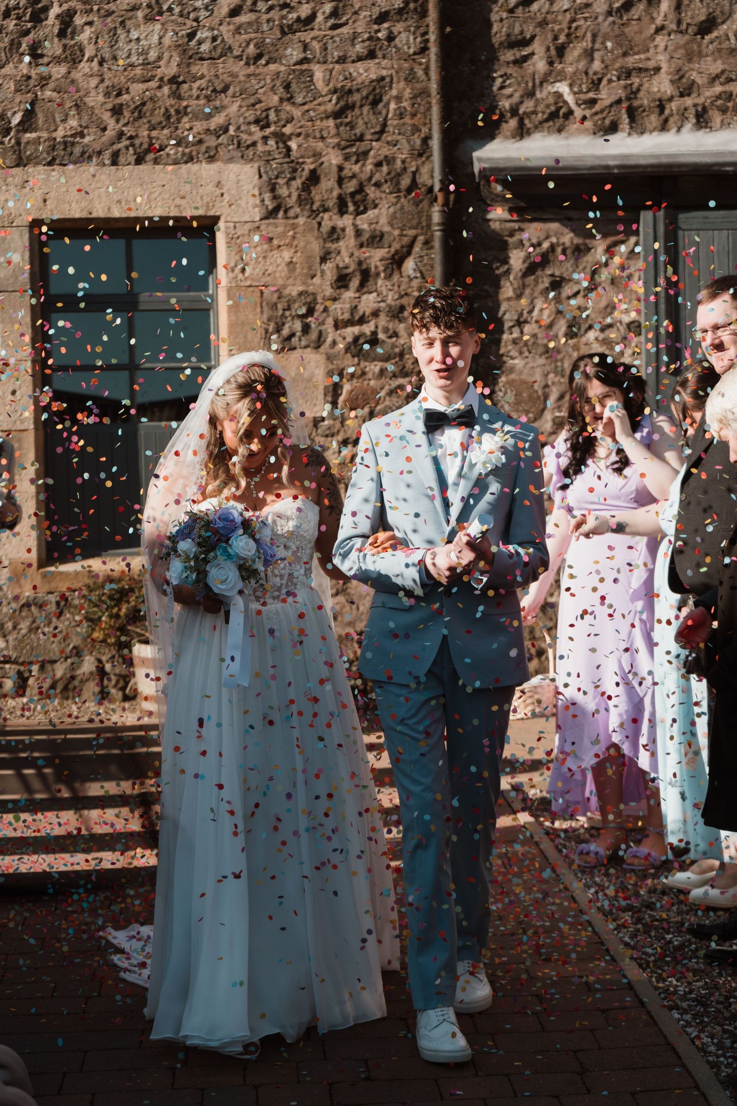 Newlywed couple walking through colorful confetti shower outside stone building, bride in white gown, groom in blue suit.