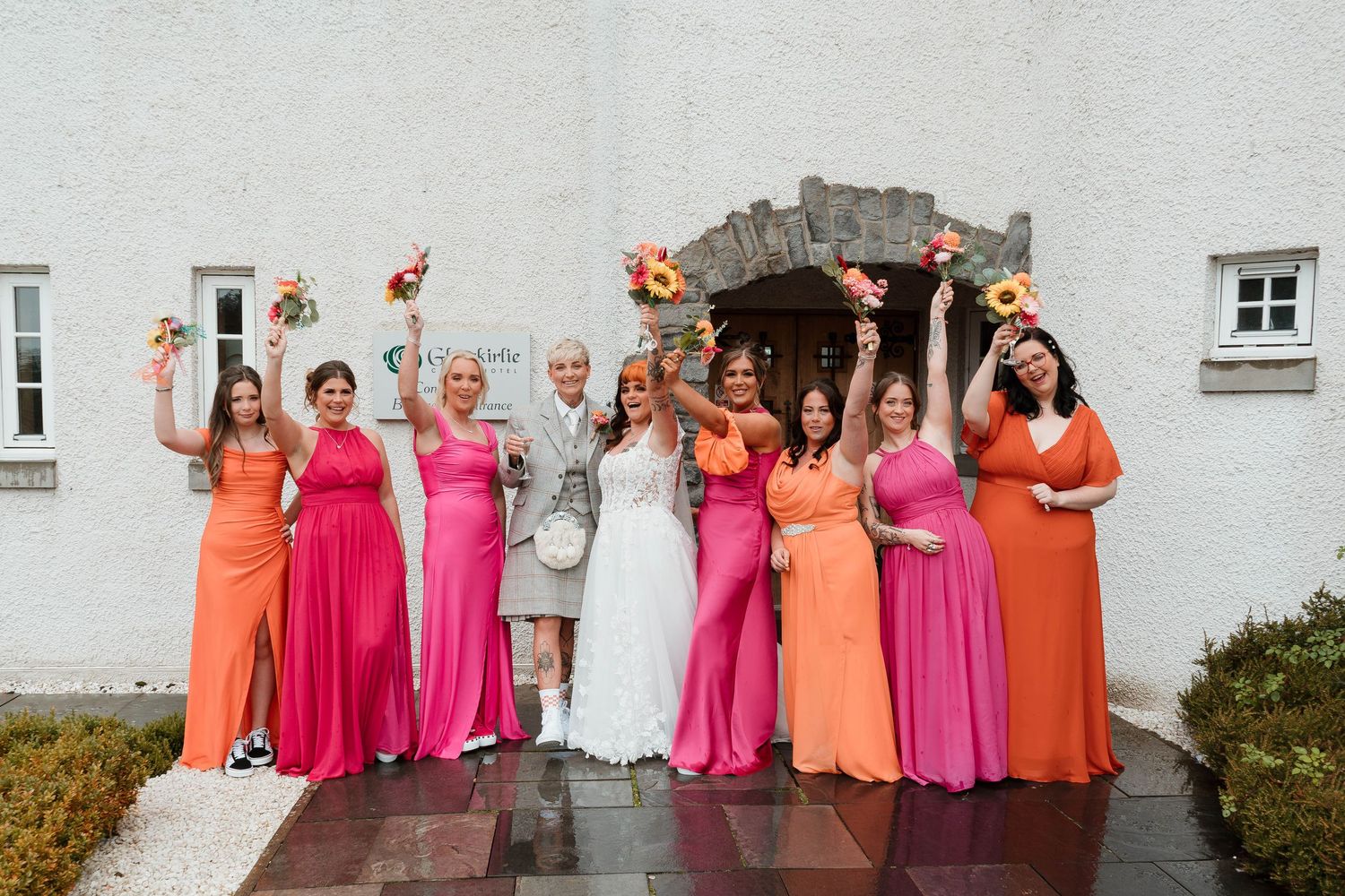 Wedding party poses against white building wearing pink and coral colored dresses.