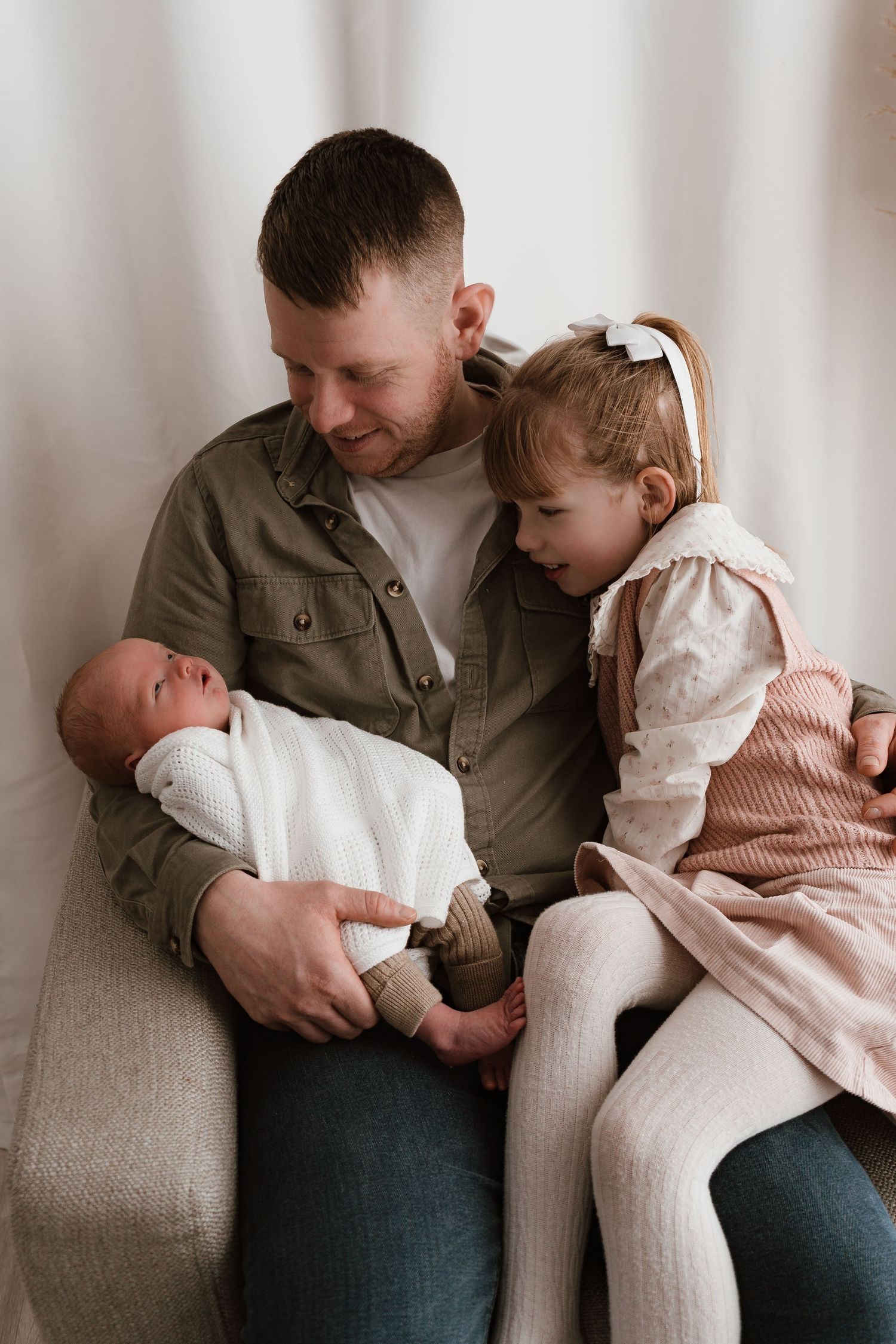 Father sitting with newborn baby and toddler daughter in white bow, sharing a tender family moment.