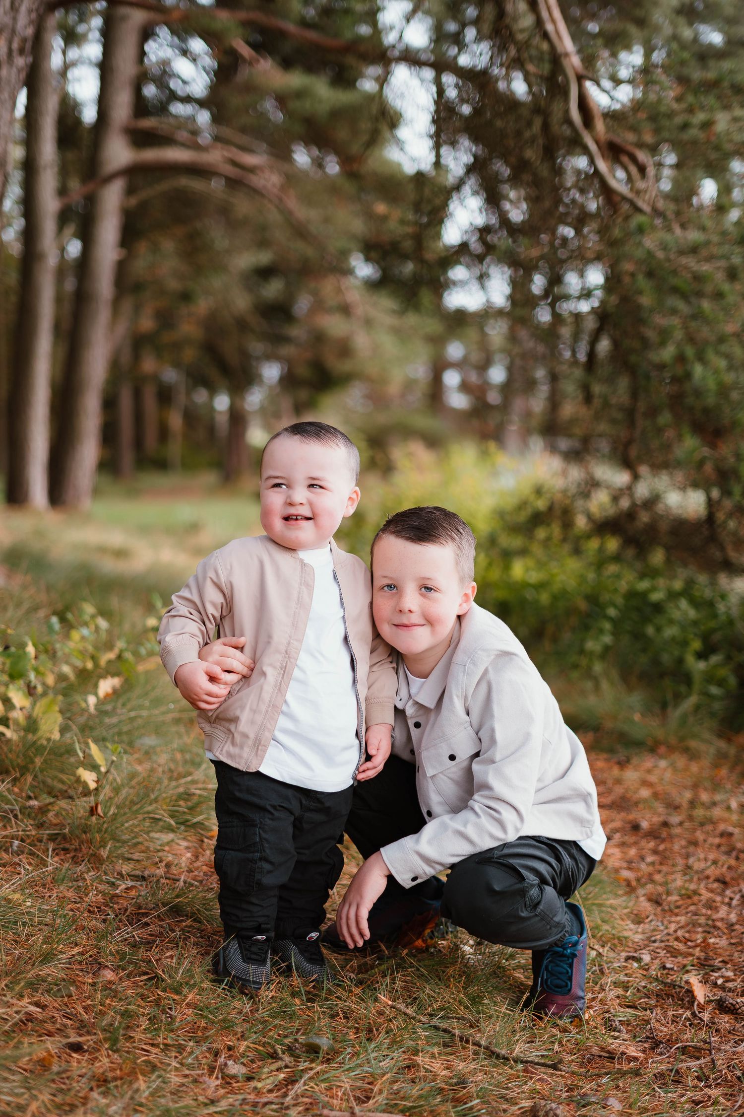 Two children pose together on fallen leaves in a wooded forest setting.