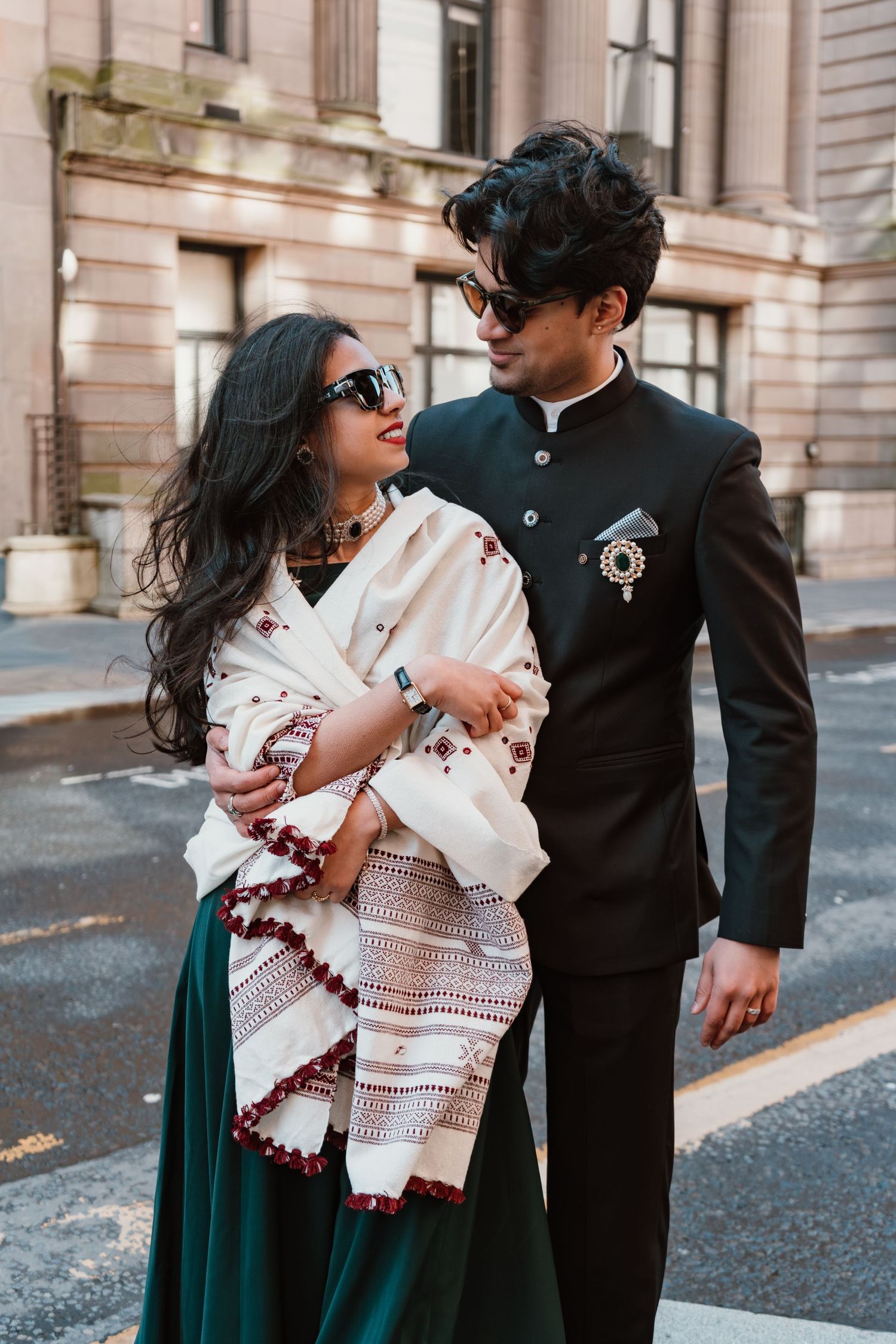 Couple in traditional Indian attire walking on city street, woman in white shawl and green skirt, man in black sherwani.