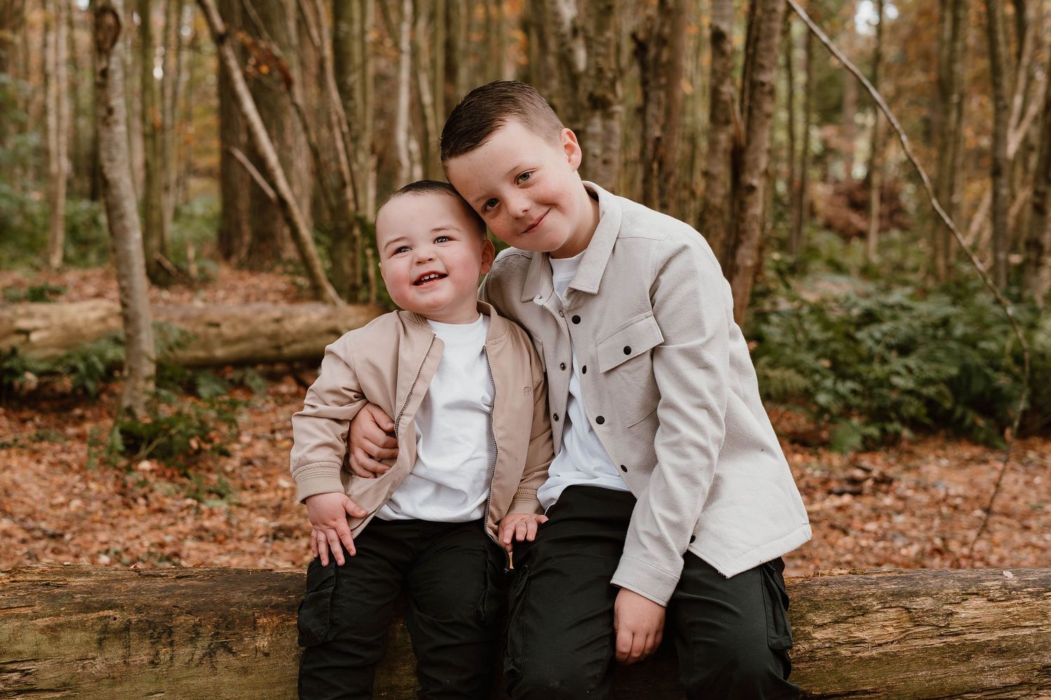Two young children in matching light-colored jackets sitting together in an autumn forest.