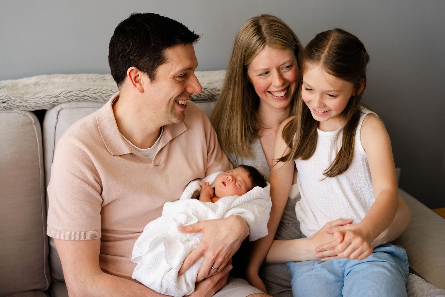 A family sits together on a couch while holding a newborn baby wrapped in white.