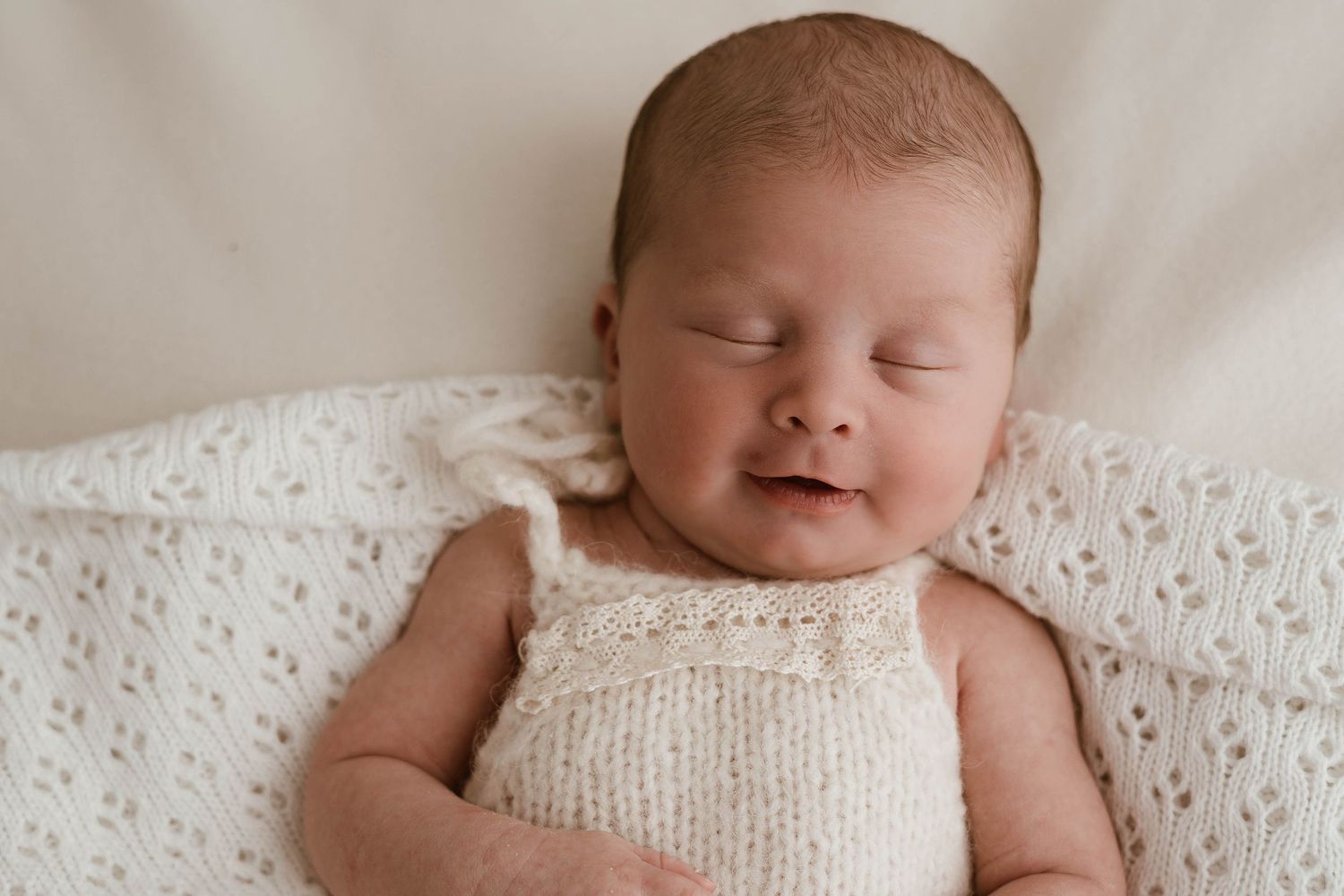 Peaceful newborn baby sleeping in a cream knit romper on a soft white blanket, smiling softly.