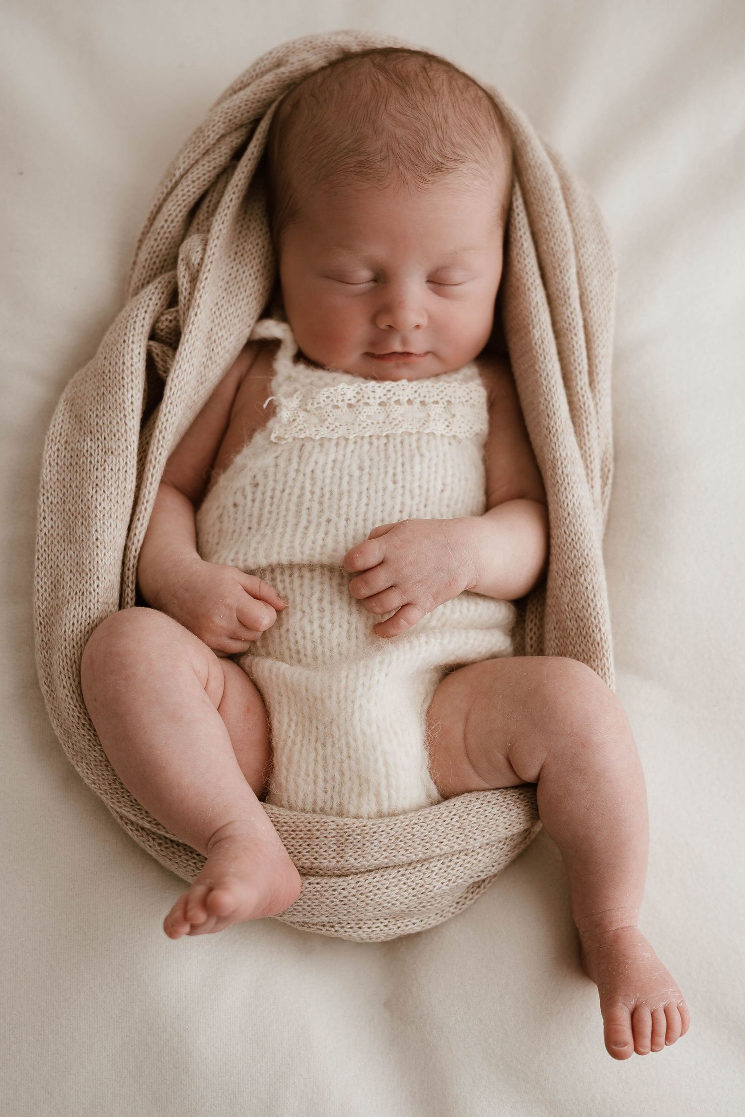 Peaceful newborn baby sleeping in a beige knit romper, wrapped in a soft tan blanket on a cream background.
