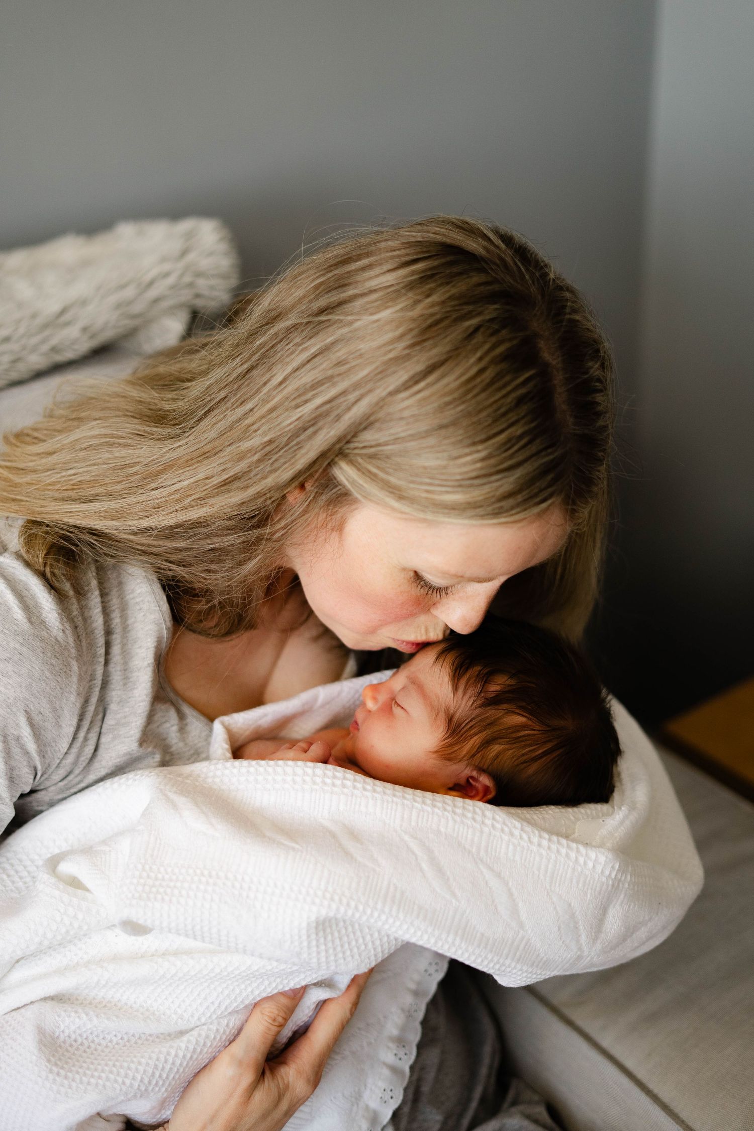 A tender moment between parent and newborn baby wrapped in white blanket while resting on grey couch.
