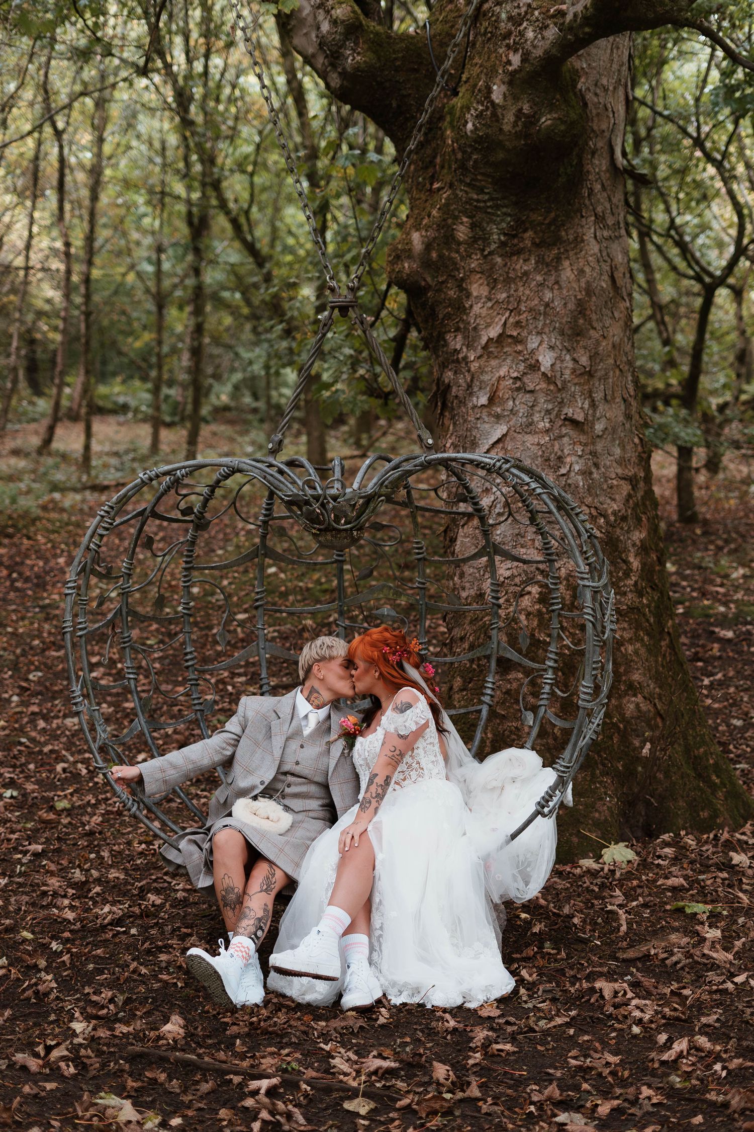 Bride and groom kissing on a heart-shaped swing surrounded by trees in a woodland wedding setting.