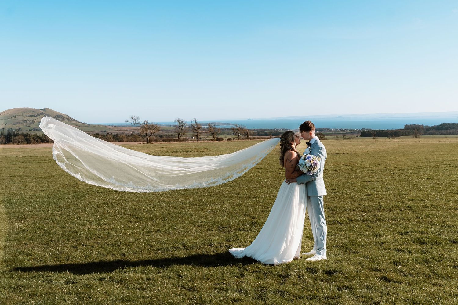 Bride and groom embrace in green field, long veil flowing in wind, scenic countryside backdrop on sunny wedding day.