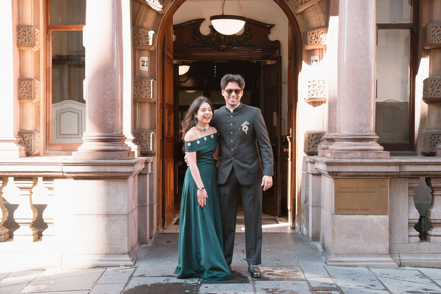 Couple posing in formal attire outside elegant stone building, woman in green gown, man in black suit.