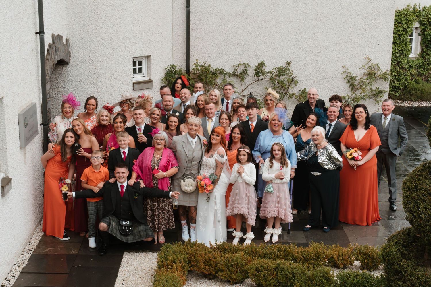 Large wedding group photo with guests lined up against white building exterior.
